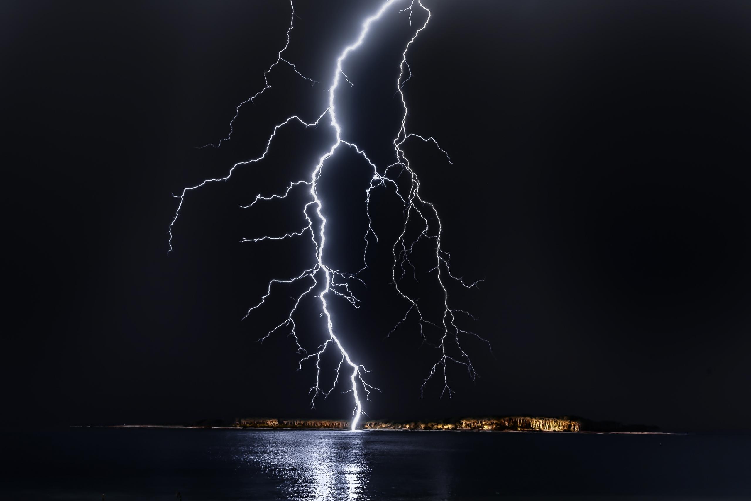 Ein heller Blitz schießt vom Himmel und schlägt vor einer Insel ins Wasser ein. 