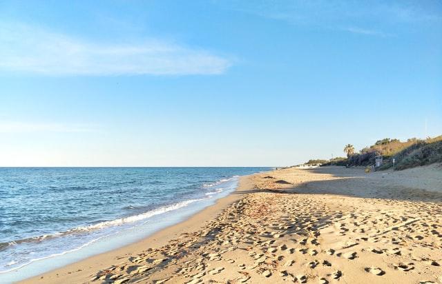 Ein heller Sandstrand mit ruhigen Wellen und Fußspuren und niedrigen Büschen am Rand passen zum ionischen Meer.