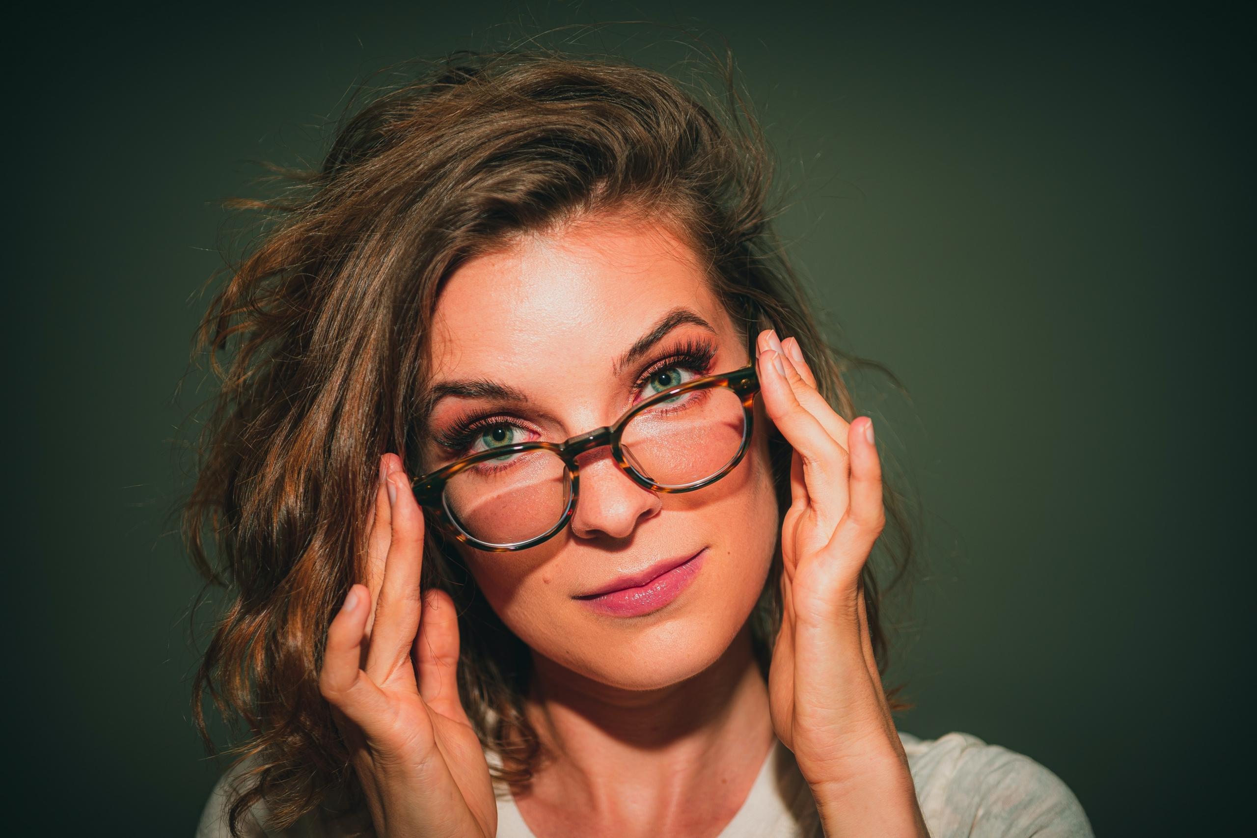 Eine Frau mit langem Haar hält den Kopf leicht geneigt und fasst mit deinen Händen an ihre Brille.