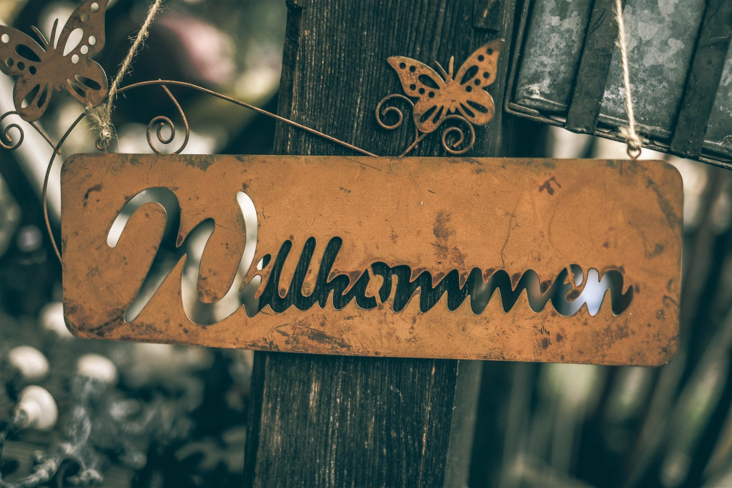 An einem Holzpfahl hängt ein Schild aus Metall mit dem Wort Willkommen.
