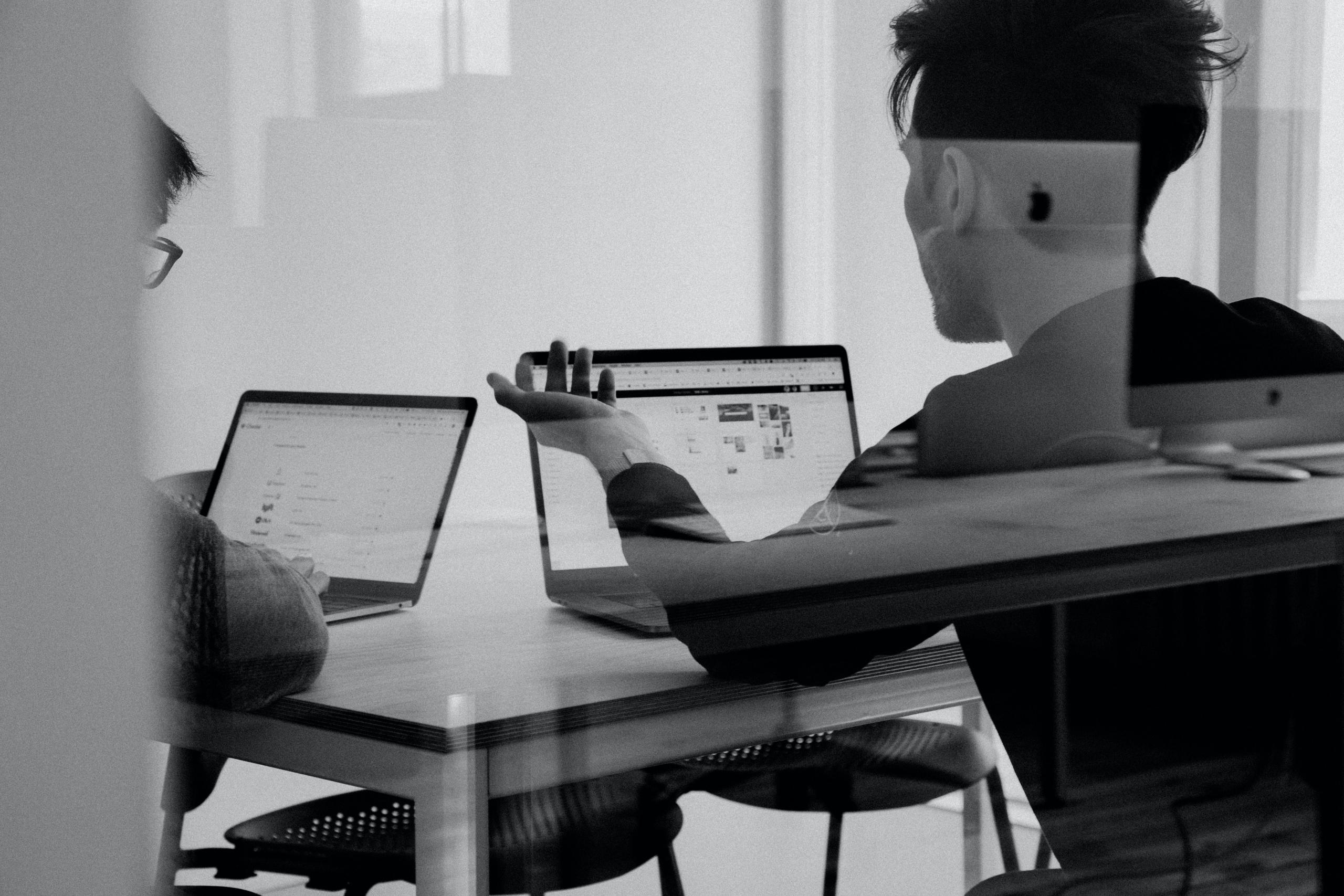 Zwei Männer sitzen in Büroraum hinter Galsscheibe vor ihren aufgeklappten Laptops und tauschen sich aus,