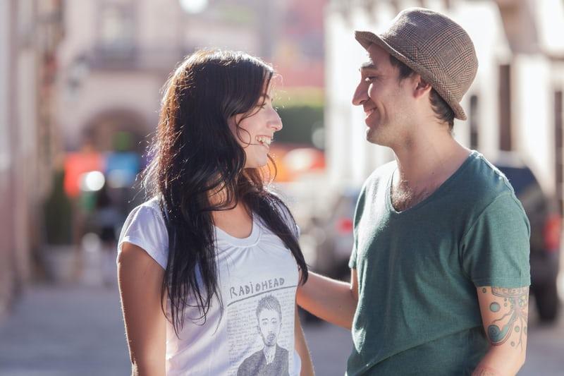 Auf einer Strasse steht ein Paar, ein junger Mann mit Hut und und eine junge Frau mit langen dunklen Haaren, die sich lächeln ansehen.