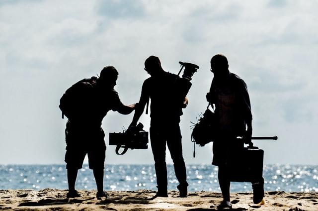 Im Gegenlicht steht eine dreiköpfige Crew für einen Film am Strand mit dem Meer im Hintergrund und trägt Equipment das es für einen Film braucht.