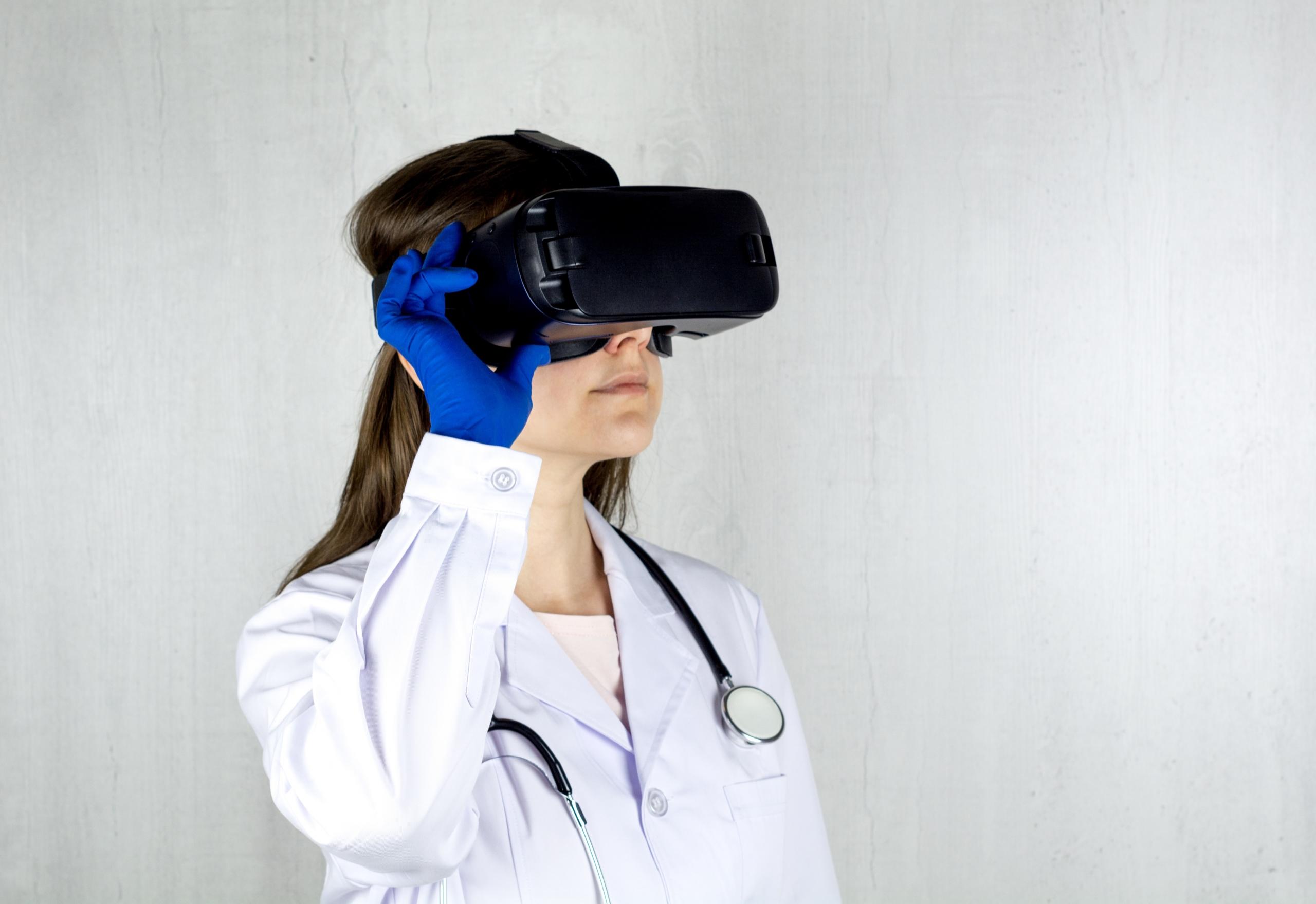 Eine Frau mit weißem Kittel und Stethoskop hat einen blauen Handschuh an und halt sich eine VR-Brille vor die Augen.