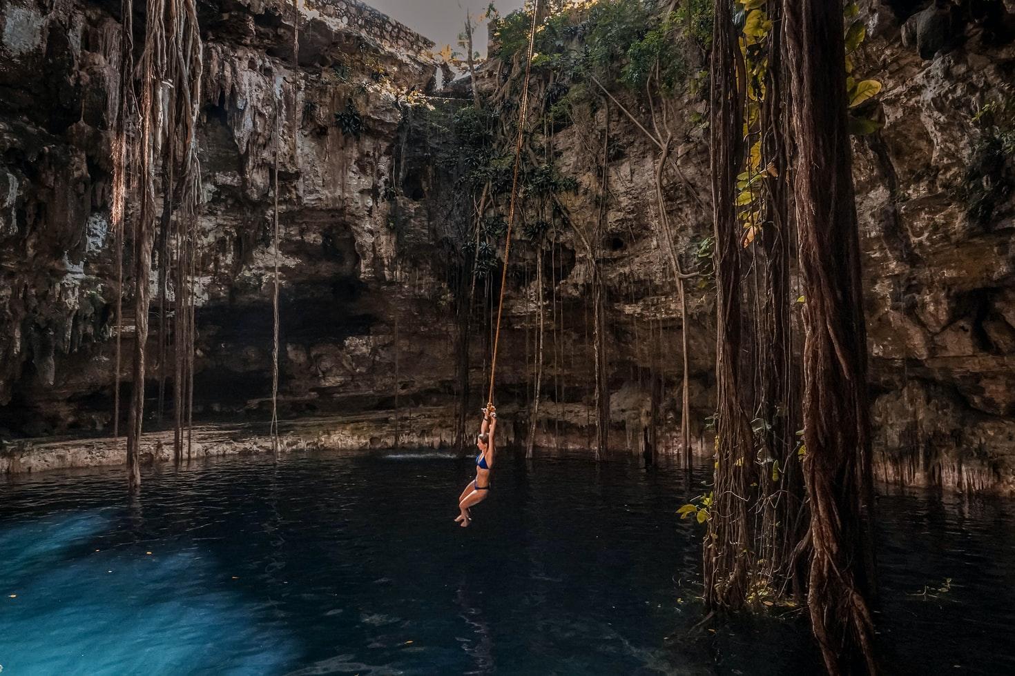 Eine Frau lässt sich über dem Wasser einer Cenote von einer Liane baumeln, die an den Felswänden der Cenote herabhängen. in eine Cenote.