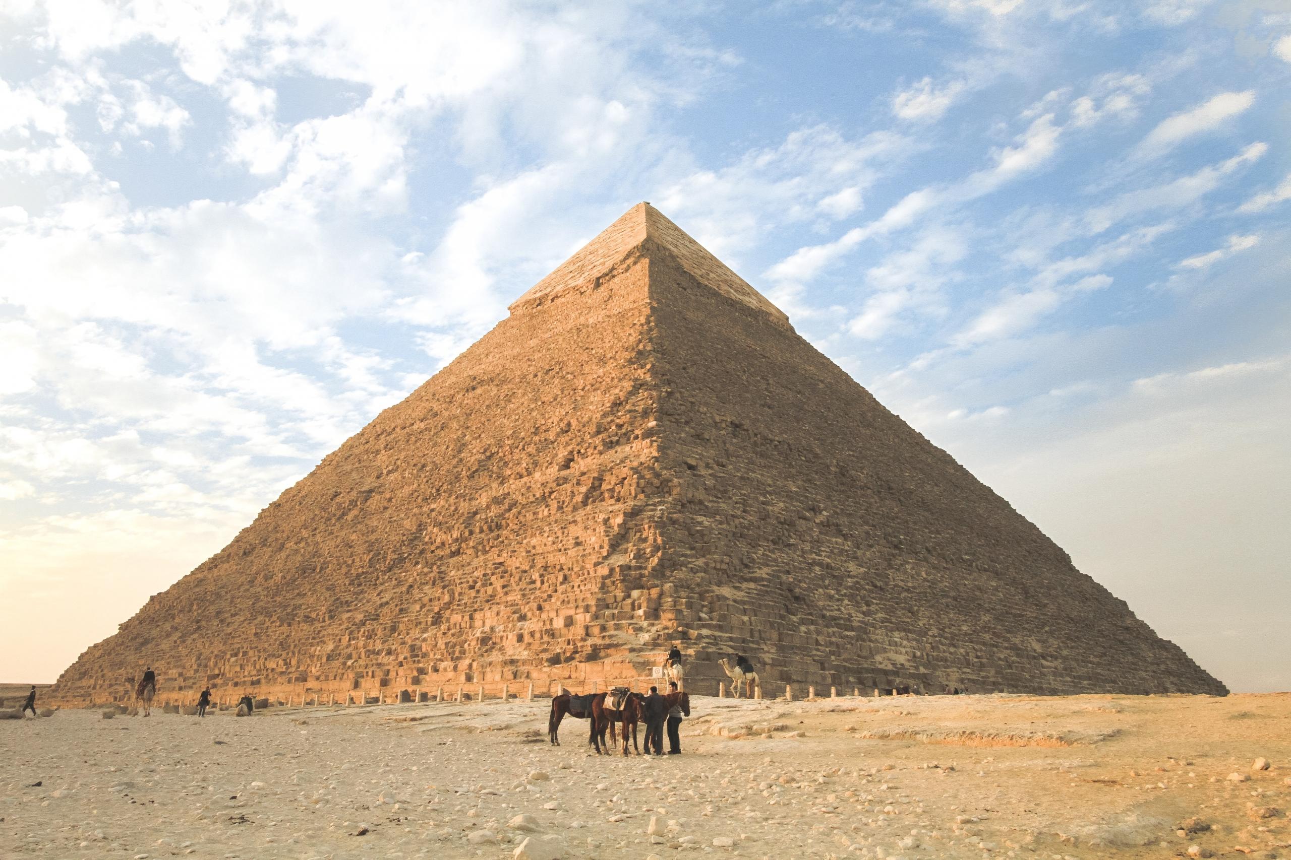 Vor einem blauen Himmel steht eine Pyramide und davor sind zwei Männer mit zwei Pferden.
