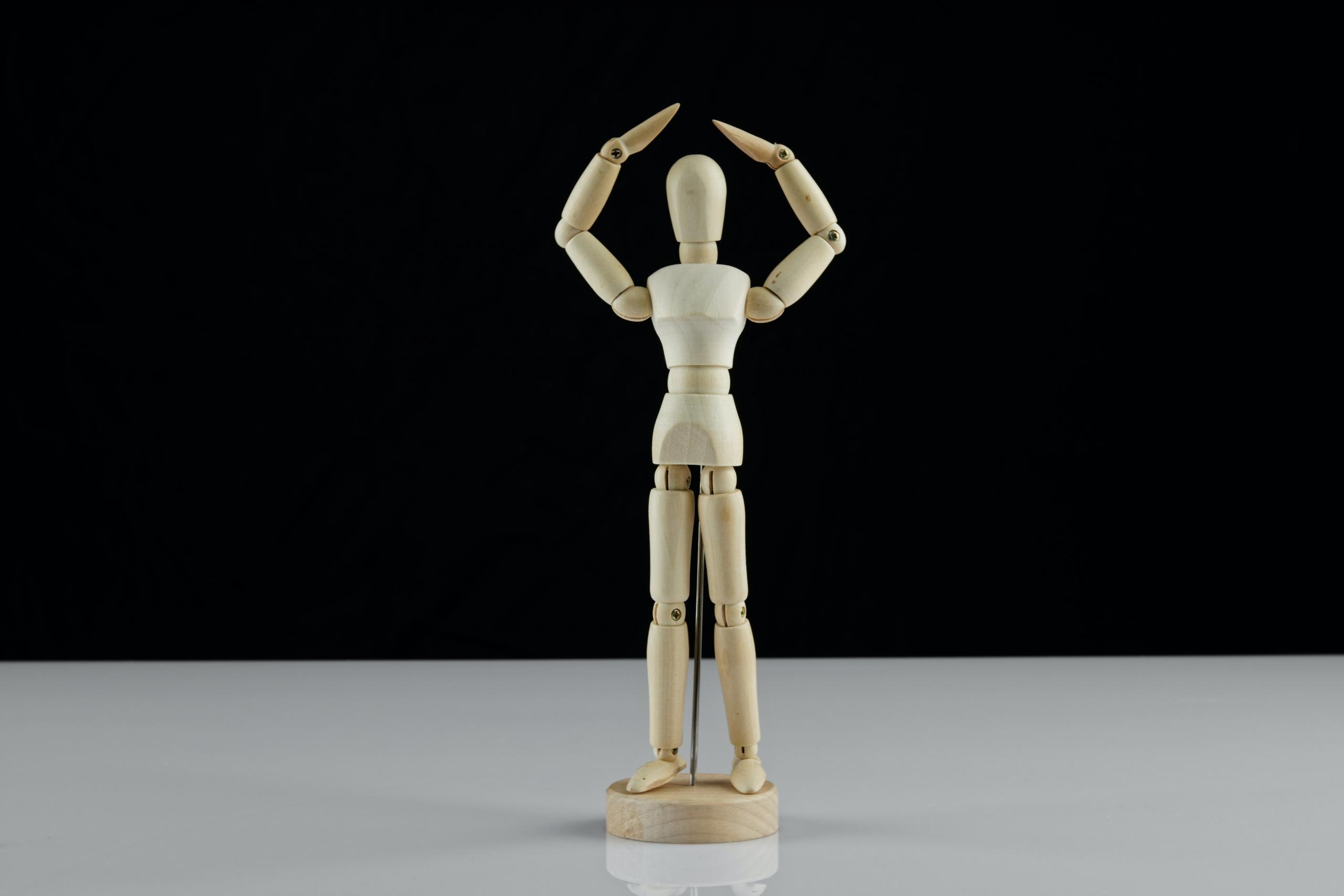 Eine Holzfigur steht ganz gerade und führt die Arme über dem Kopf zusammen.