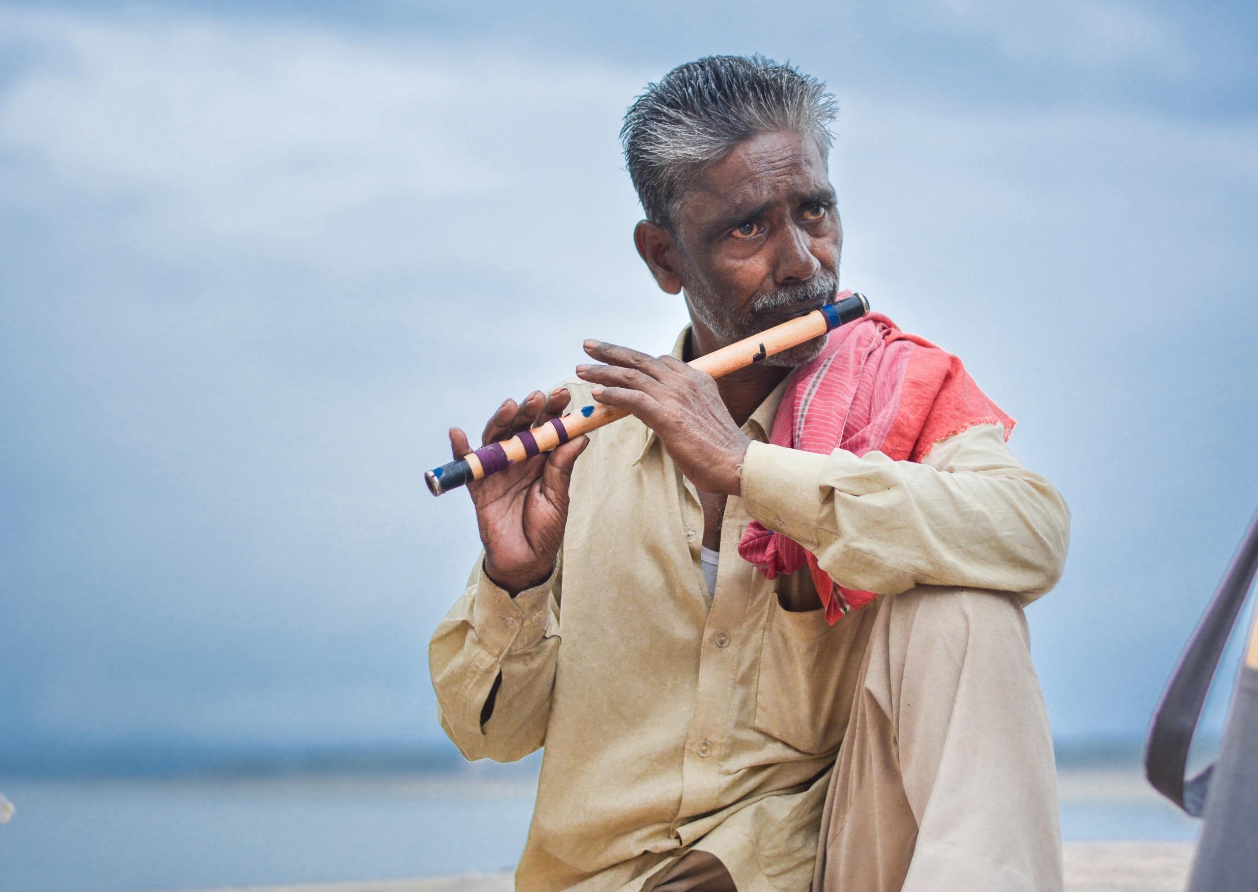Ein Mann mit grauen Haaren spielt vor blauem Himmel eine traditionelle indische Flöte.