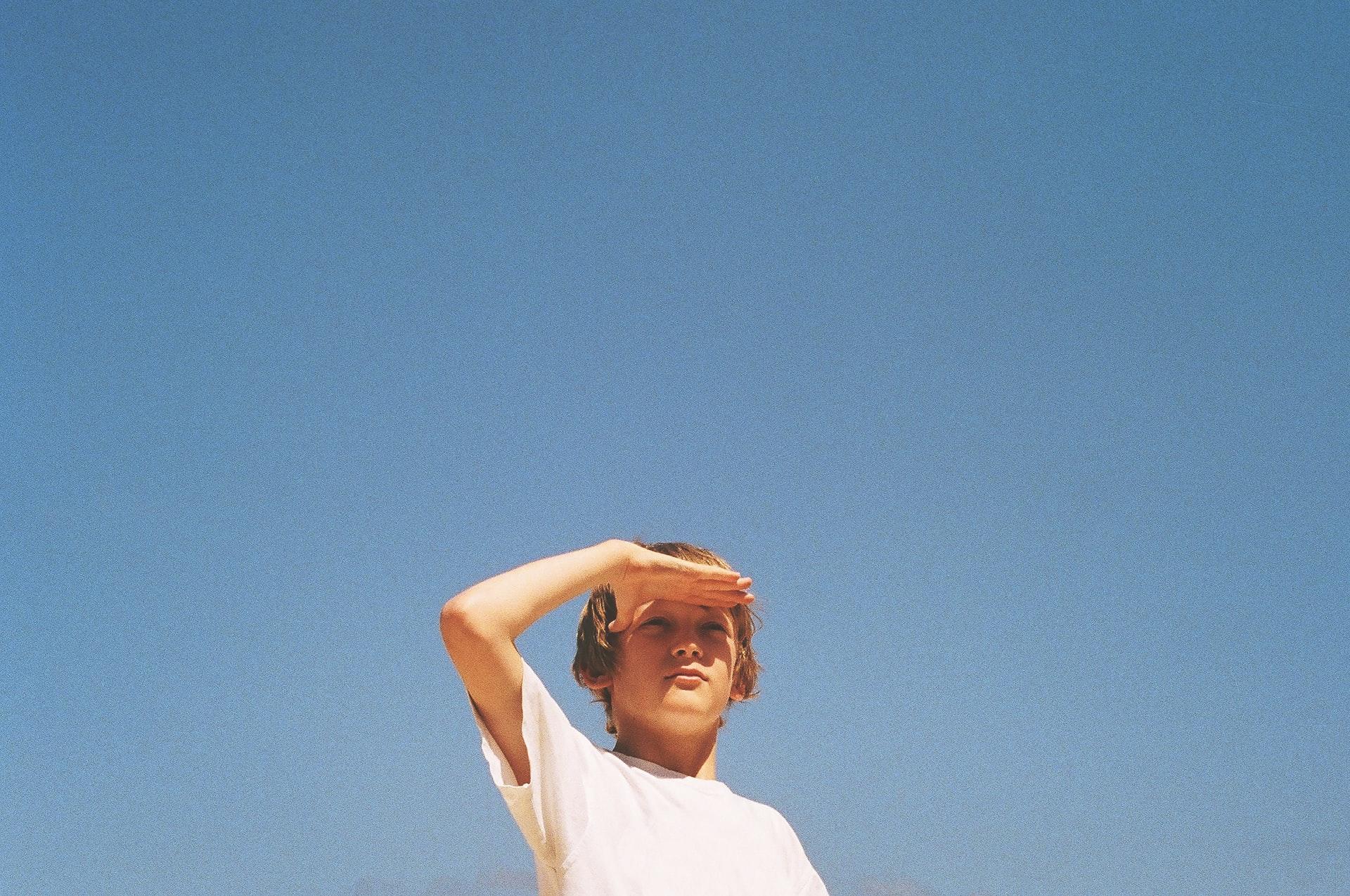 Ein Kind steht vor dem blauen Himmel und schaut mit an die Stirn gelegter Hand in die Ferne.