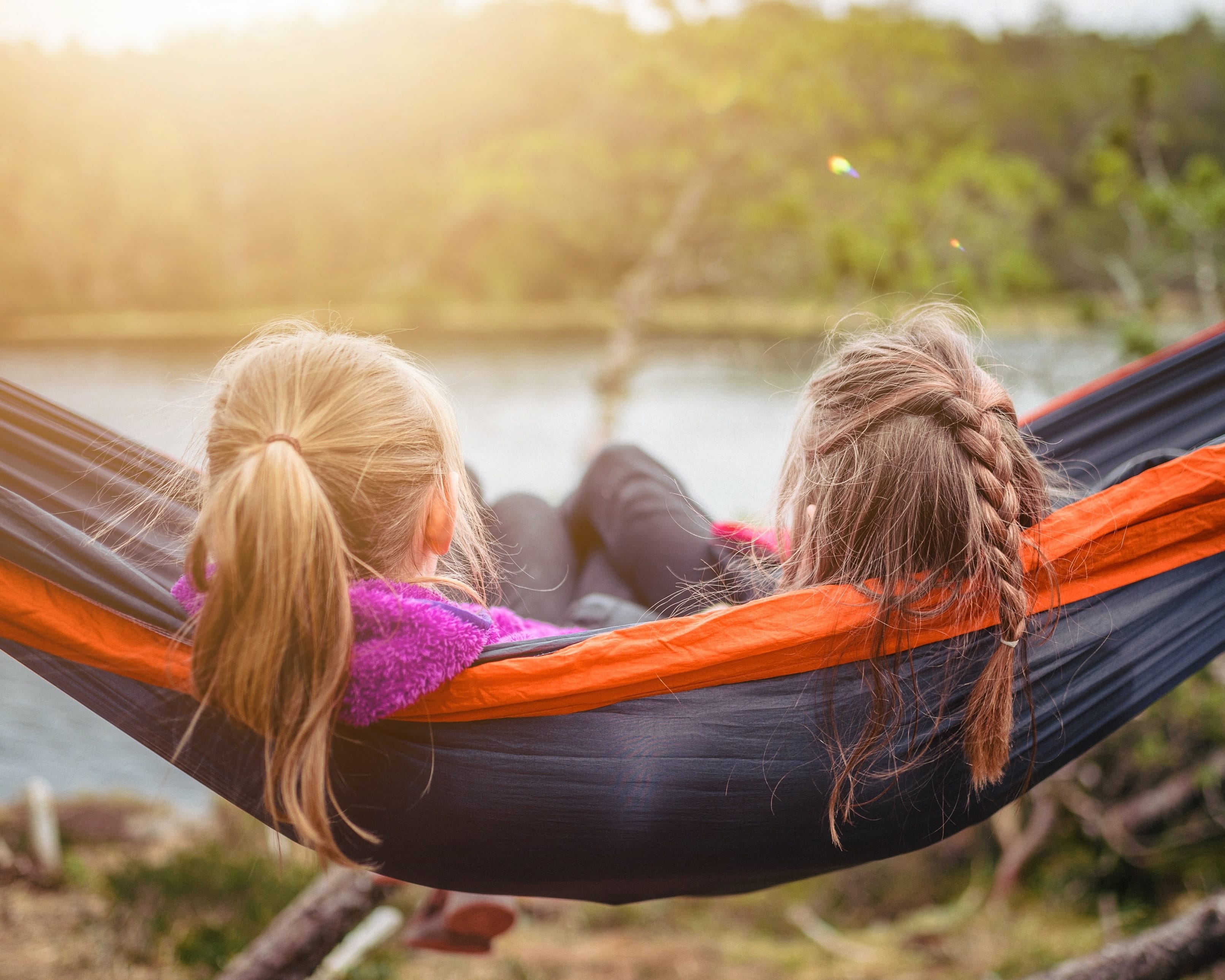 Zwei Kinder mit langen Zöpfen liegen zusammen in einer Hängematte und schauen auf einen See.