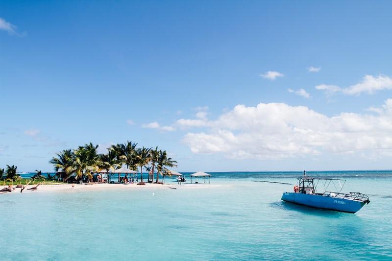 Eine kleine Insel mit weissem Sand und Palmen, umgeben von blauem Wasser, im Vordergrund ein kleines Boot.