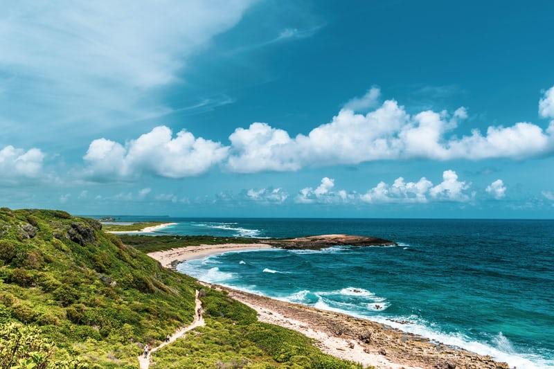 Blick auf das Meer und einen felsigen Strand von einem grünen Hügel, am blauen Himmel stehen einige weisse Wolken.