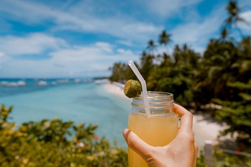 Eine Hand hält einen Rum-Cocktail, im Hintergrund Sandstrand, Palmen, blaues Wasser.