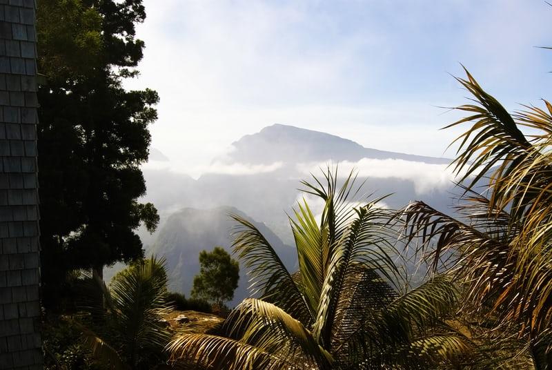 Blick über ein Tal mit weissen Wolken im blauen Himmel, in der Ferne ein weiterer Berg, im Vordergrund Palmen.