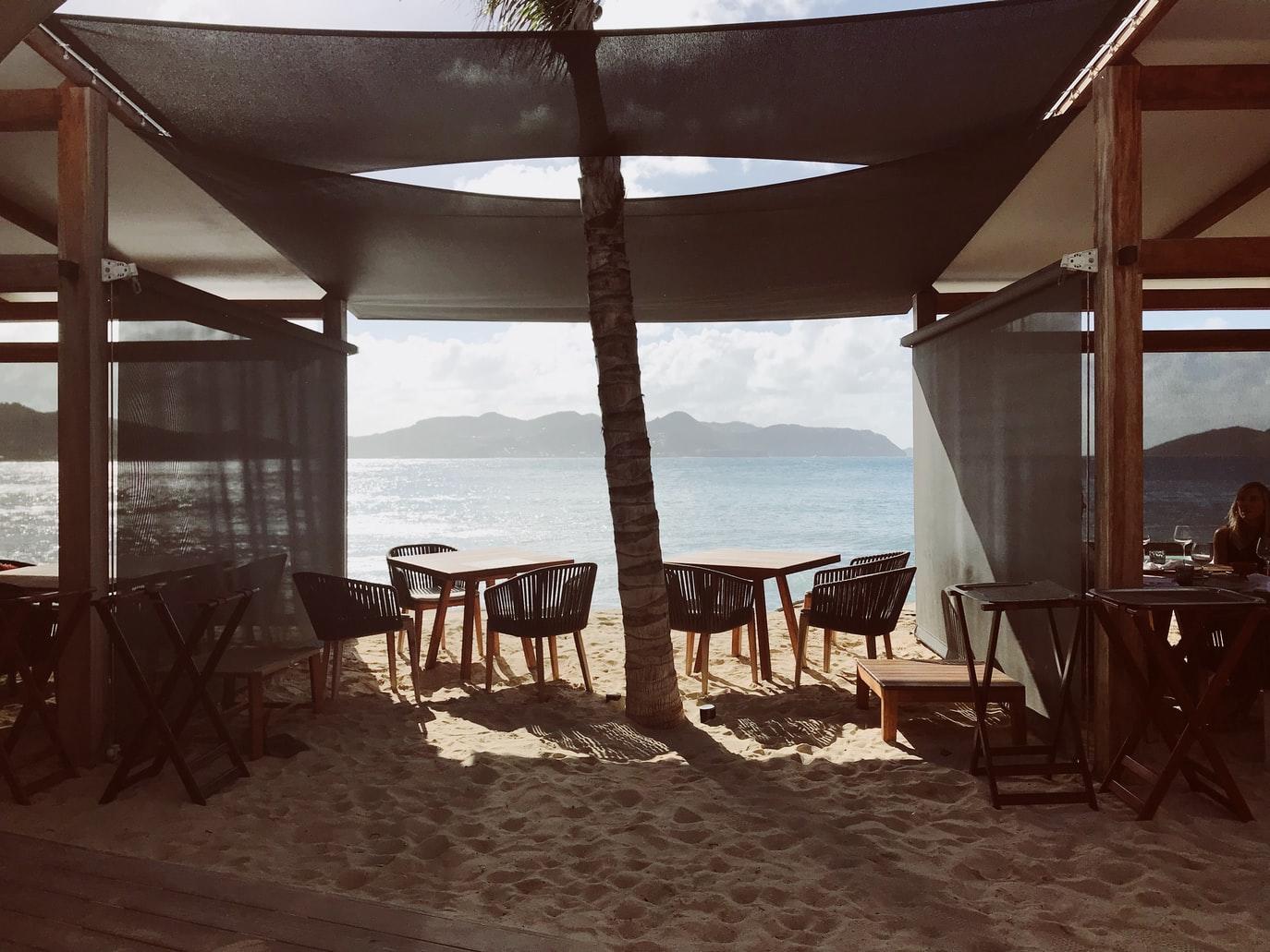 Strandbar unter Zeltdach, Blick aufs Meer, Tische und Stühle stehen im Sand.