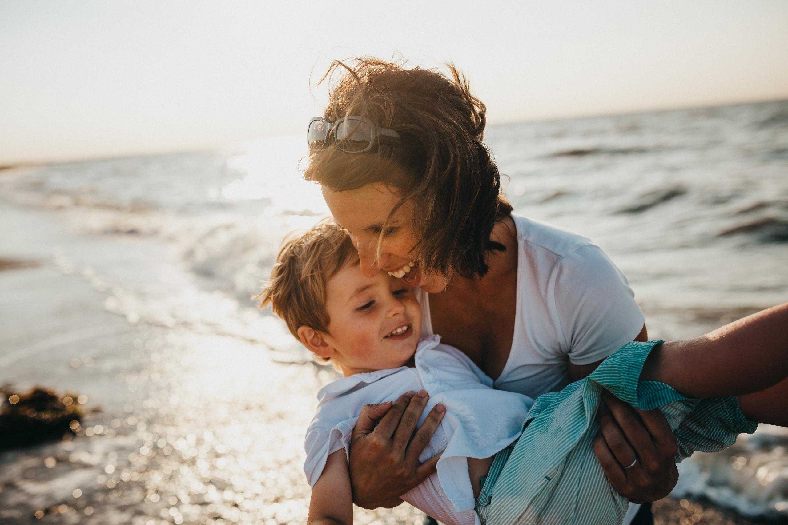 Eine lachende Mutter hält ihren Sohn am Strand auf dem Arm, im Hintergrund das Meer.
