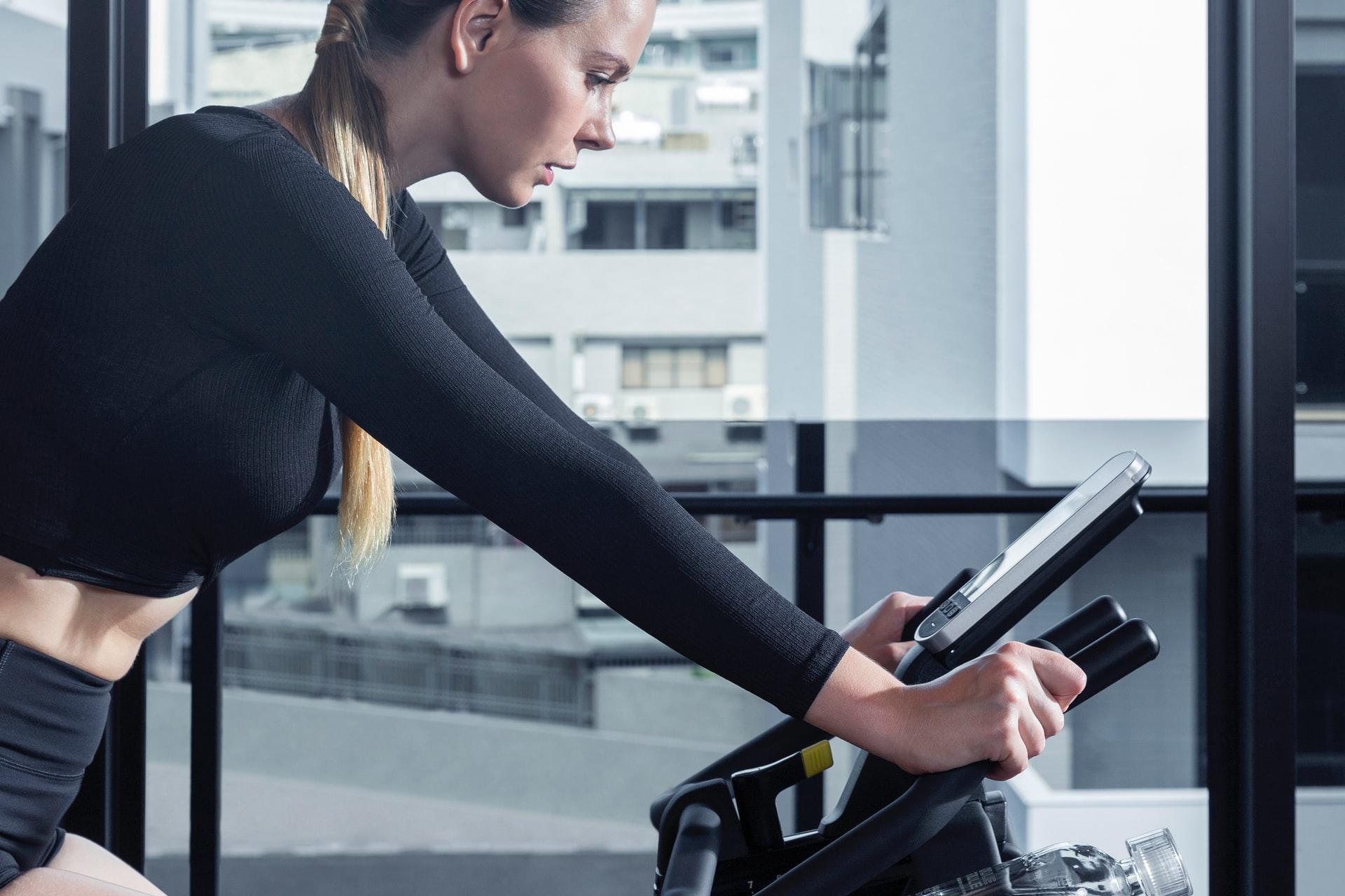 Eine junge Frau mit langem Haar sitzt konzentriert auf einem Heimtrainer und ist von der Seite fotografiert.