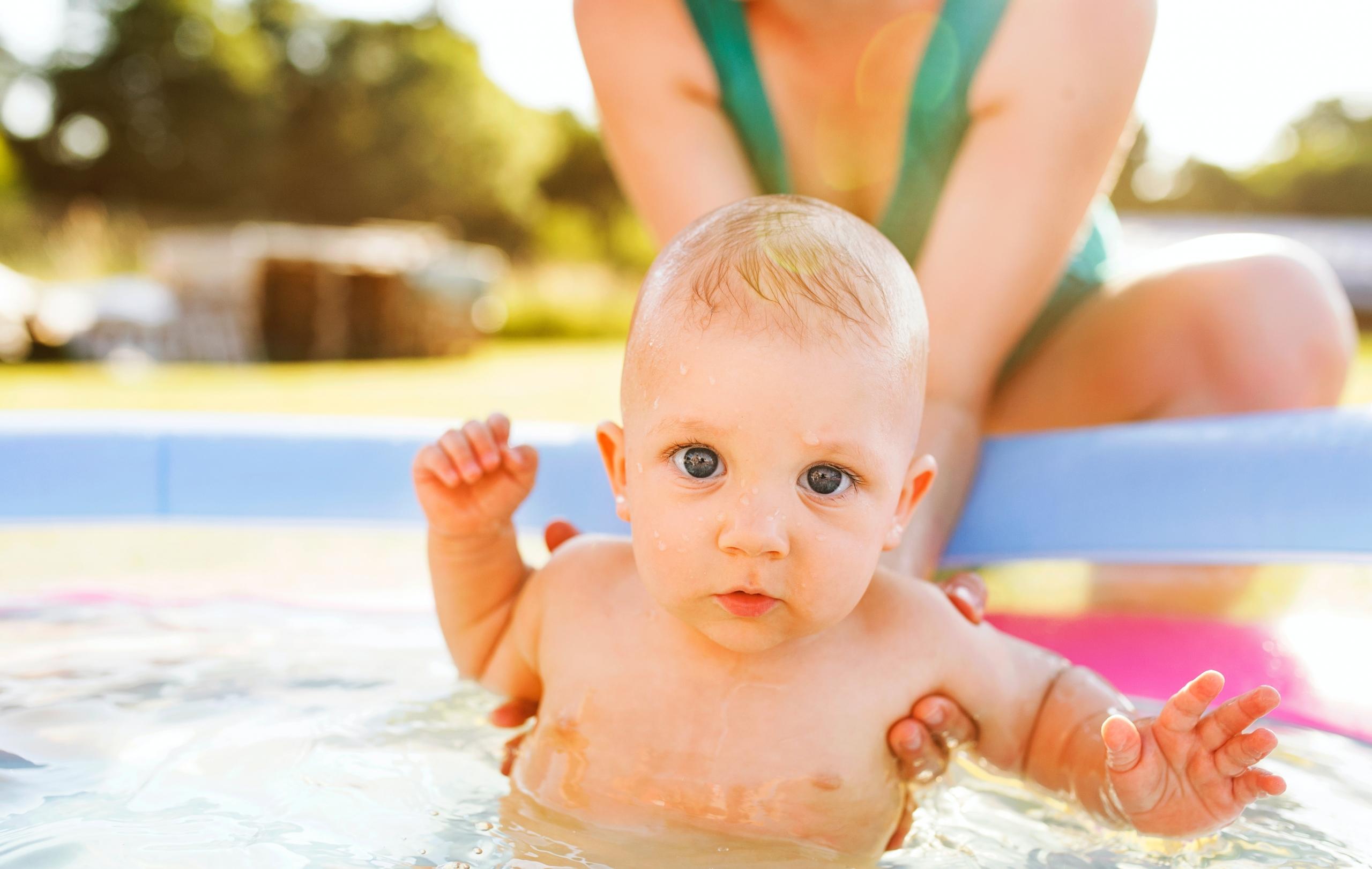 Ein Baby wird von zwei Händen im Wasser in einem Planschbecken gehalten.