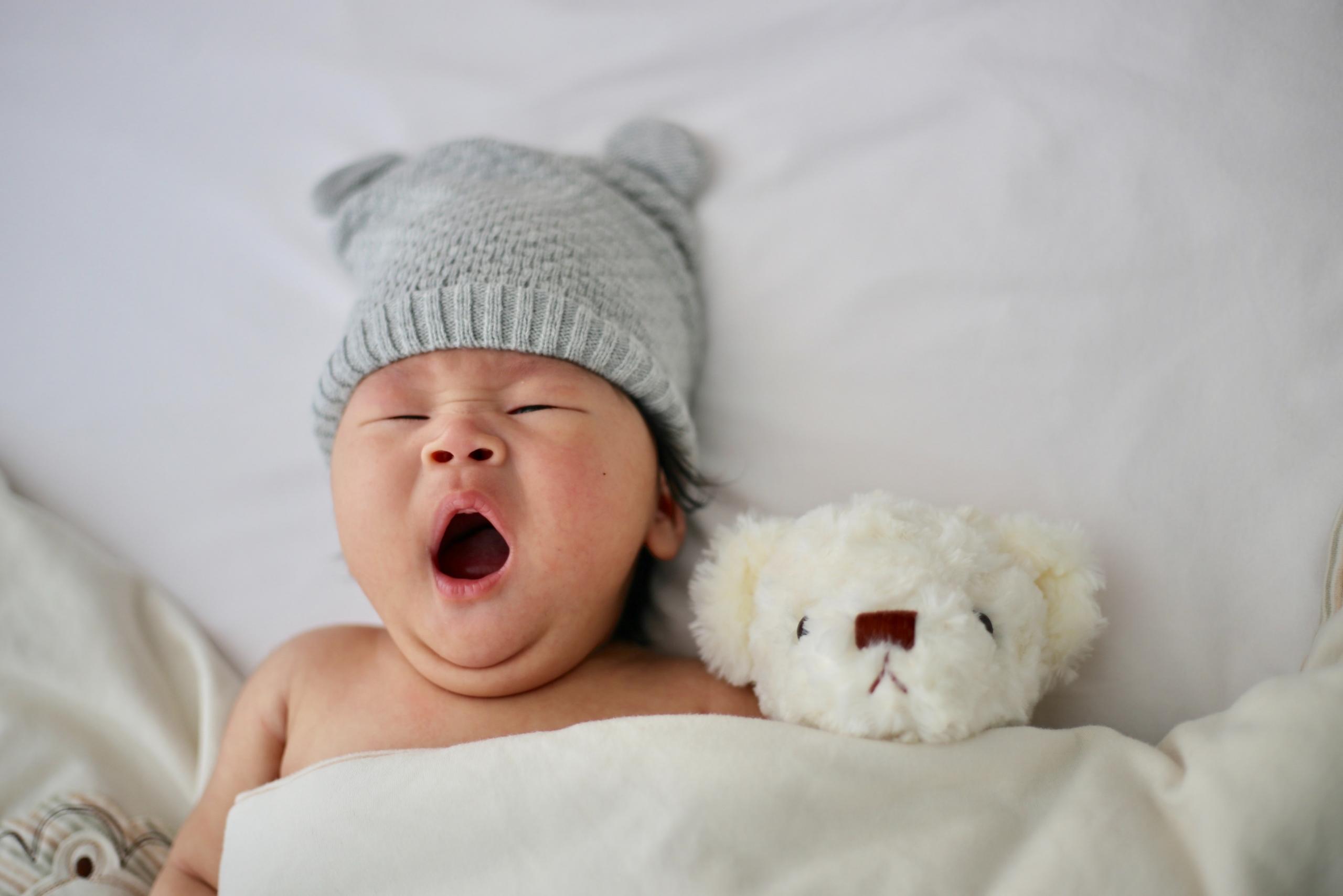Auf einem weissen Laken liegt ein gähnendes Baby mit einer Mütze auf dem Kopf unter einer weissen Decke. Neben ihm liegt ein weisser Teddybär.