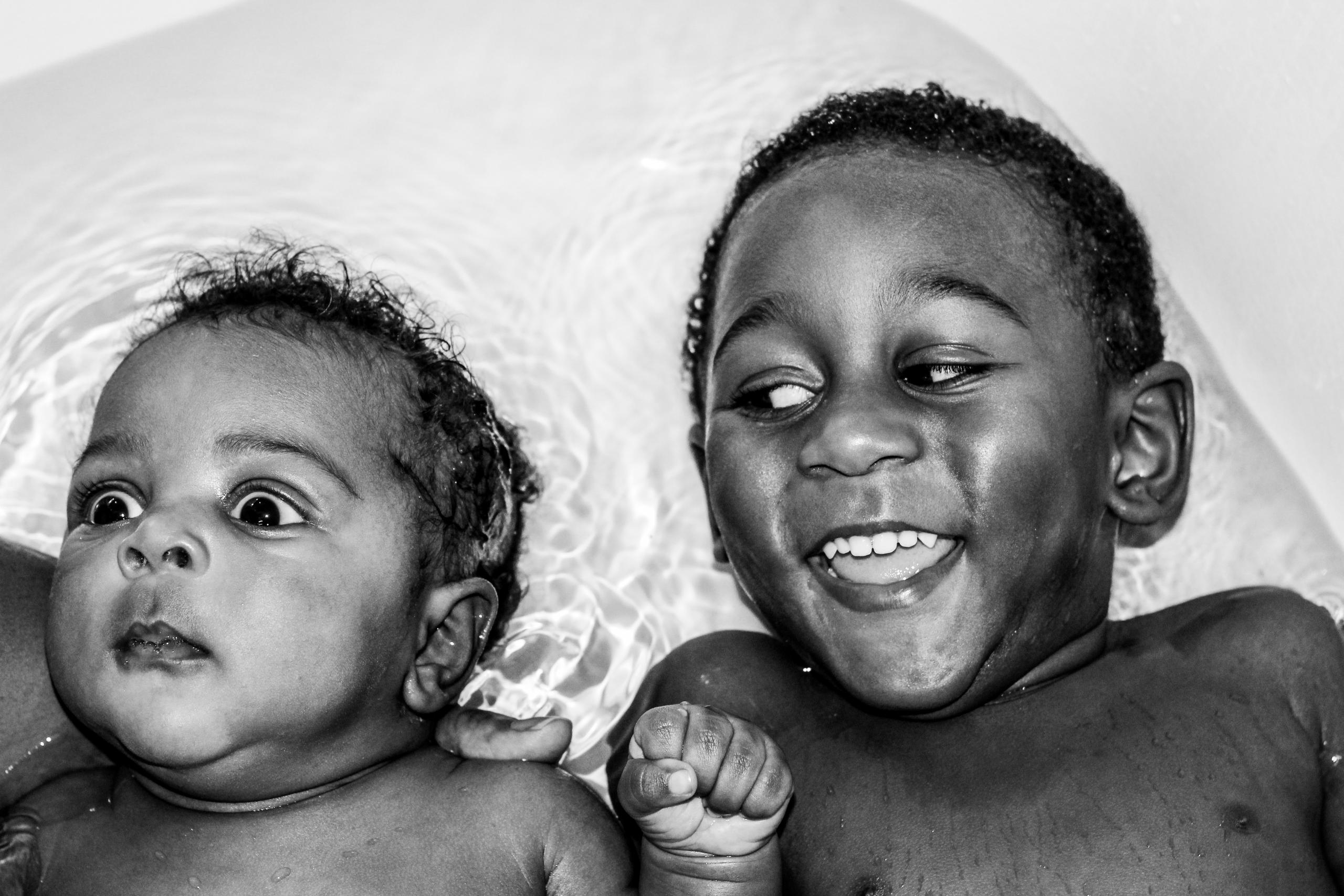 In einer Badewanne wird ein Baby von einem Erwachsenen gehalten. Sein grosser Bruder lacht über sein schockiertes Gesicht.