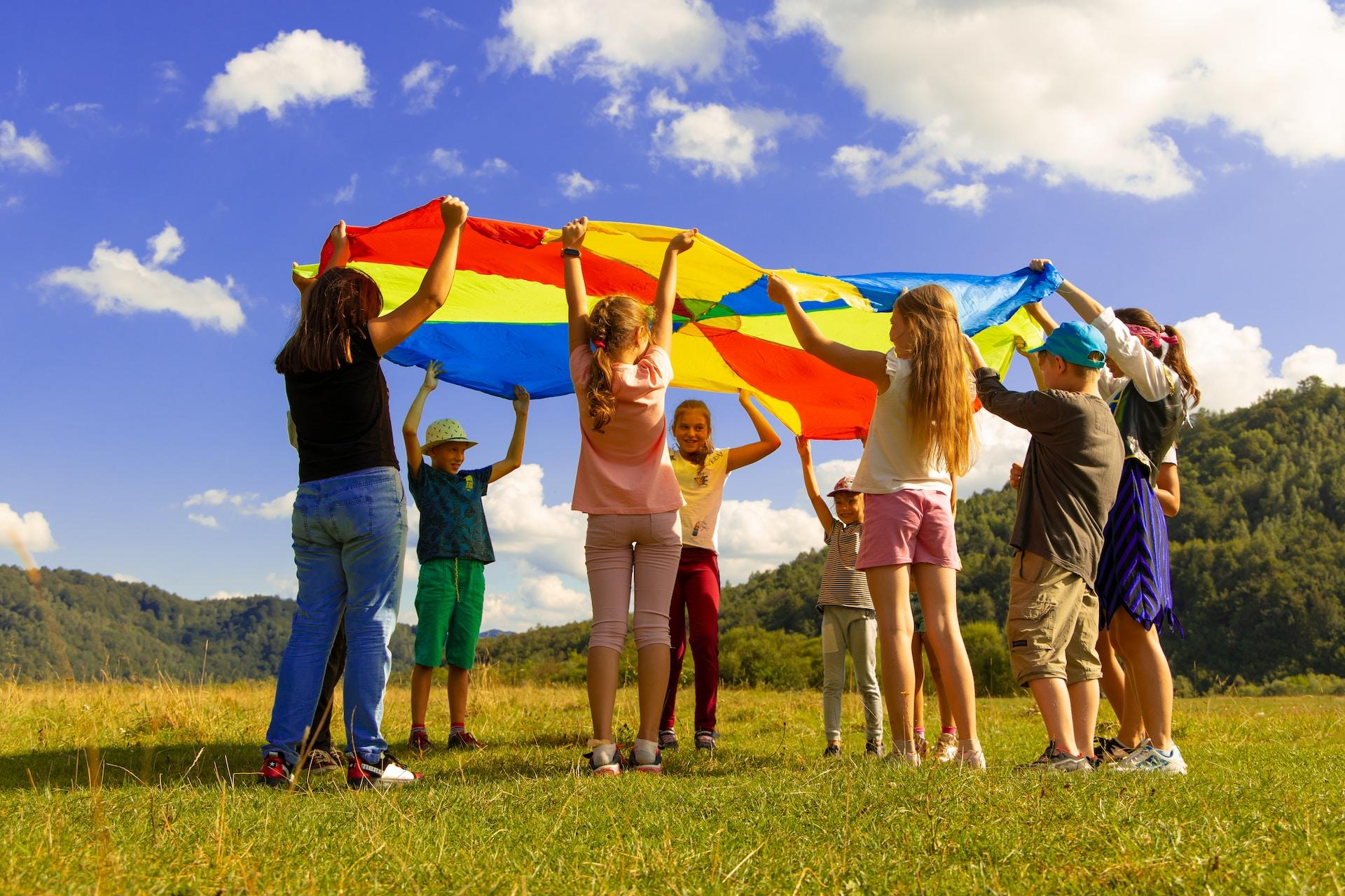 Kinder stehen auf einer Wiese unter blauem Himmel im Kreis und spielen mit einem alten Fallschirm.