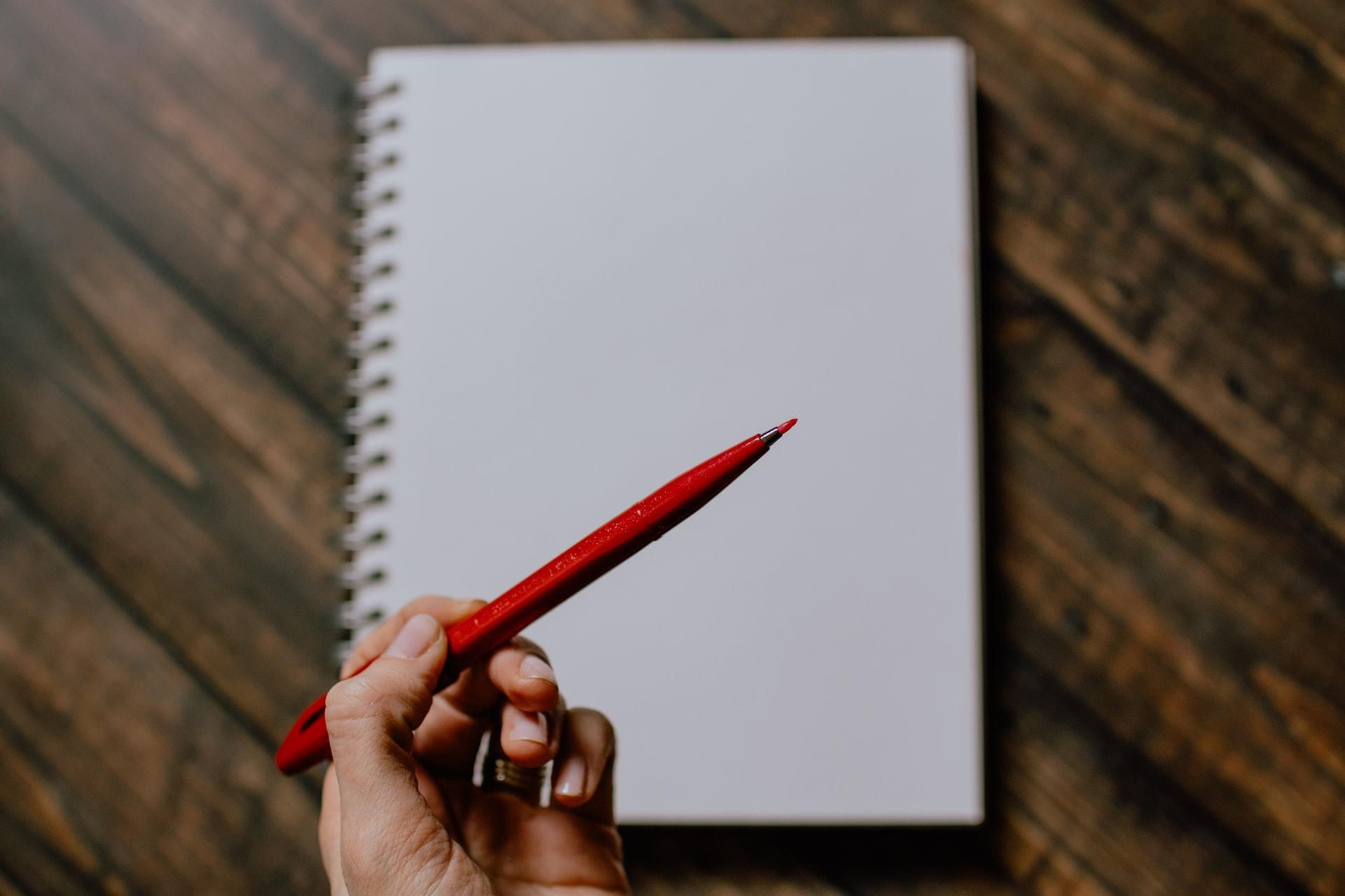 Eine Hand hält einen roten Stift über einem leeres Notizblatt in die Höhe.