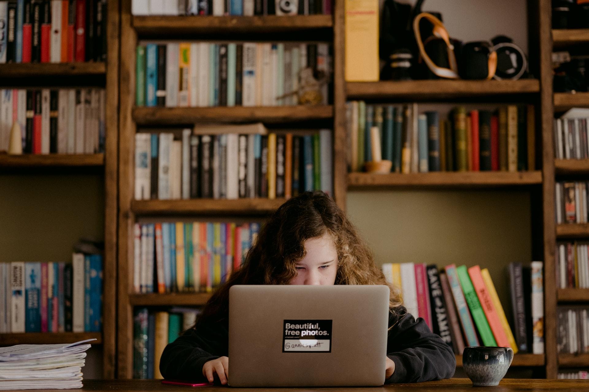 Eine junge Frau in schwarzem Pullover sitzt vor einer Bücherwand an ihrem Laptop.