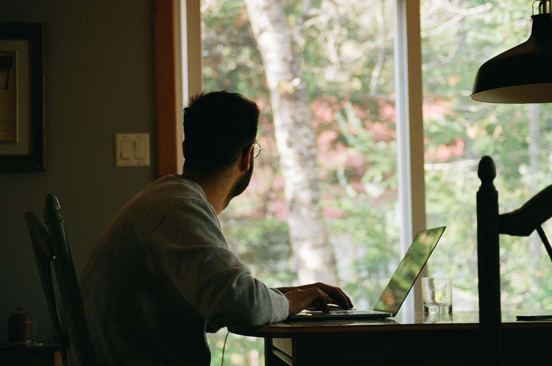 Ein Mann sitzt an seinem Laptop und blickt durch ein Fenster.