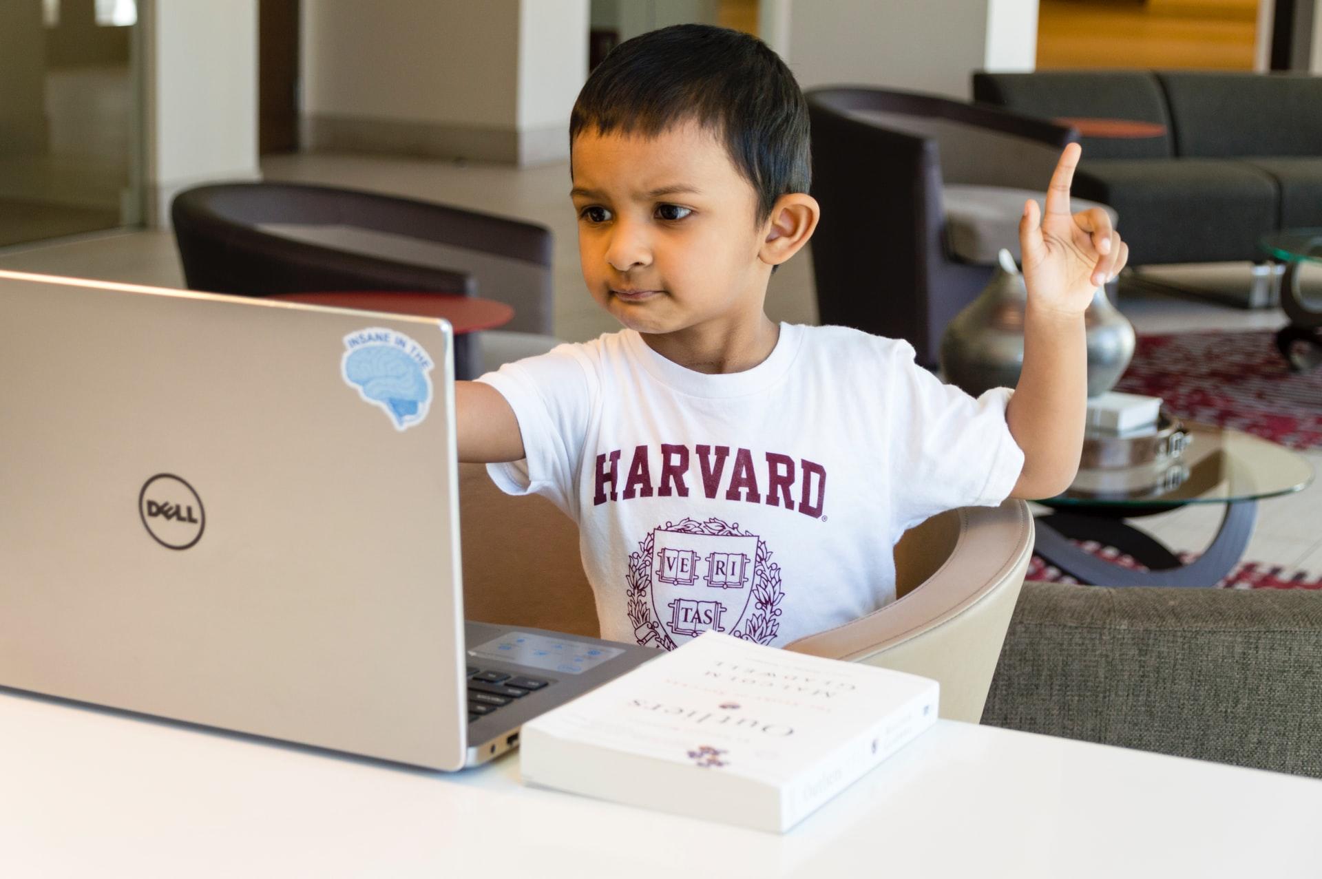 Ein Junge in weißem T-Shirt sitzt auf einem Stuhl vor einem aufgeklappten Laptop.