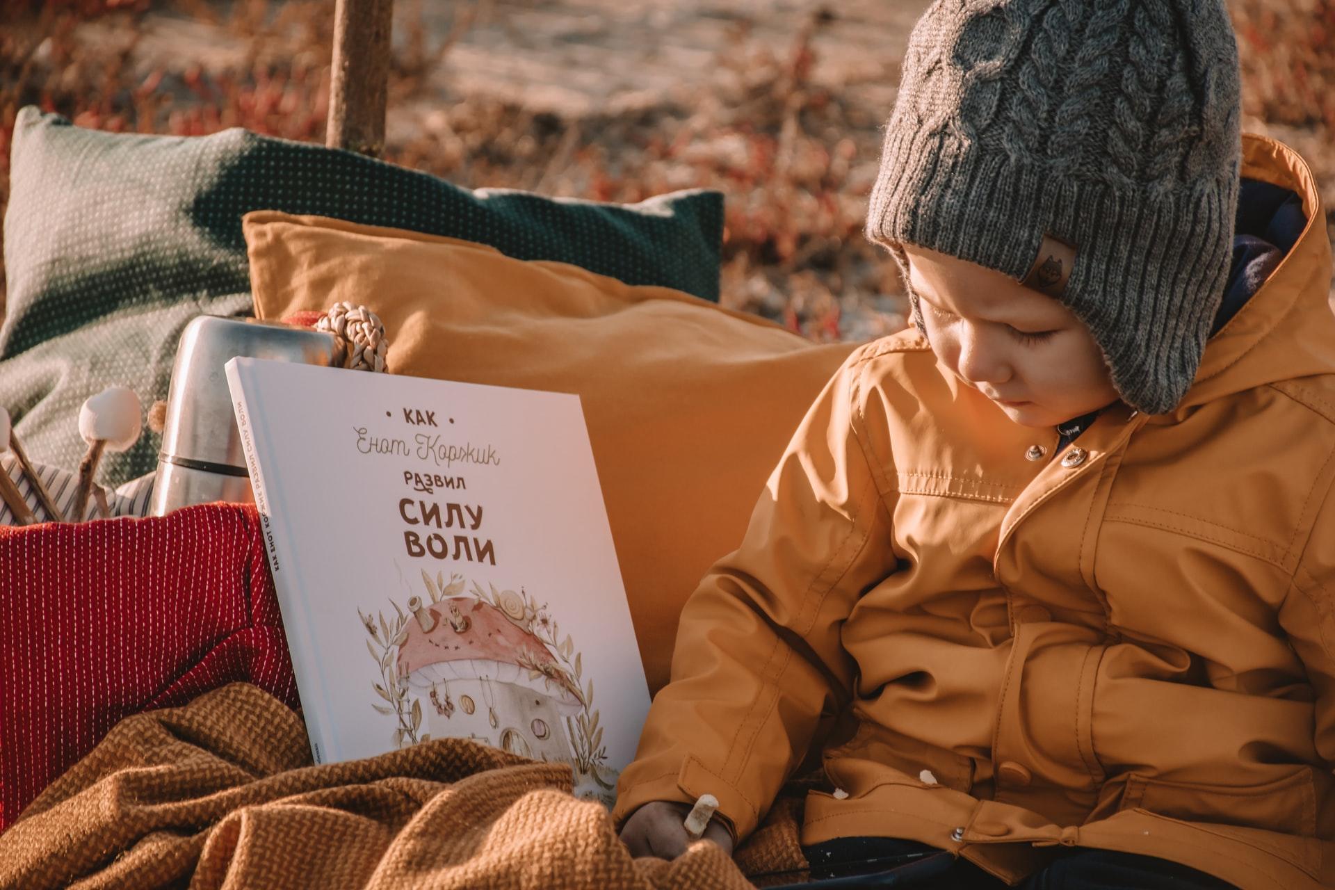 Ein russisches Kinderbuch liegt neben einem kleinen Kind mit Mütze und orangefarbener Jacke.