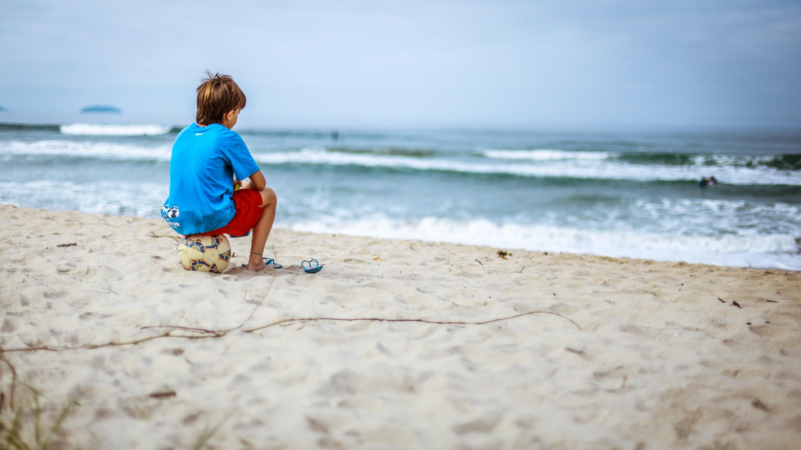 Ein kleiner Junge sitzt am Strand auf einem Fussball und schaut aufs Meer.