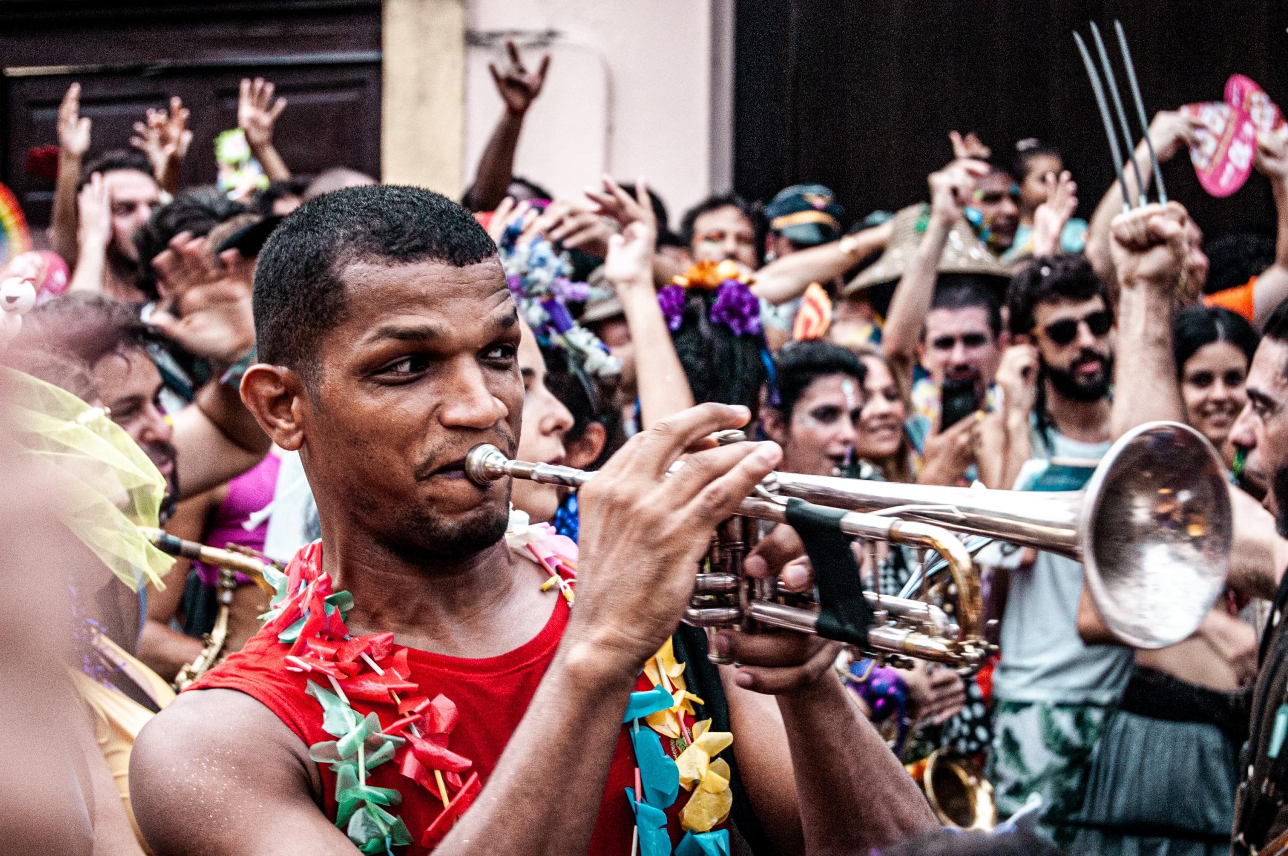 Auf einer Strassenparade spielt ein Mann mit Blumenkette inmitten jubelnder Menschen Trompete. 