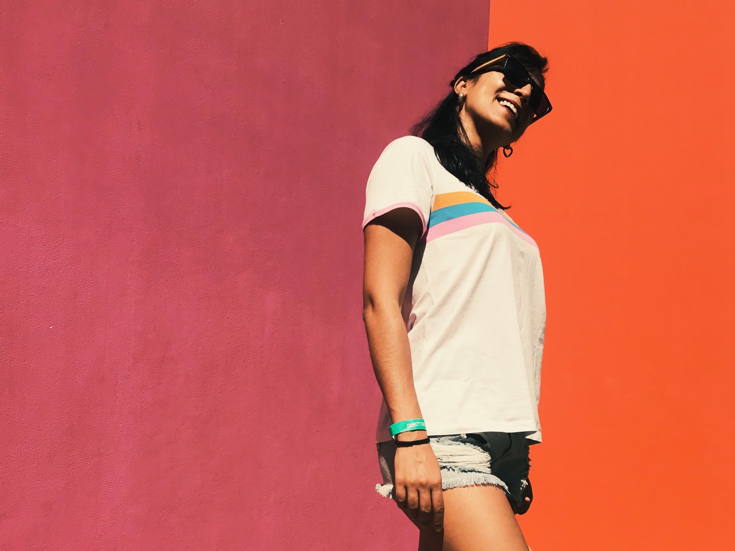 Eine dunkelhaarige Frau mit Sonnenbrille, T-Shirt und Shorts steht lächelnd vor einer pink-orangenen Wand.
