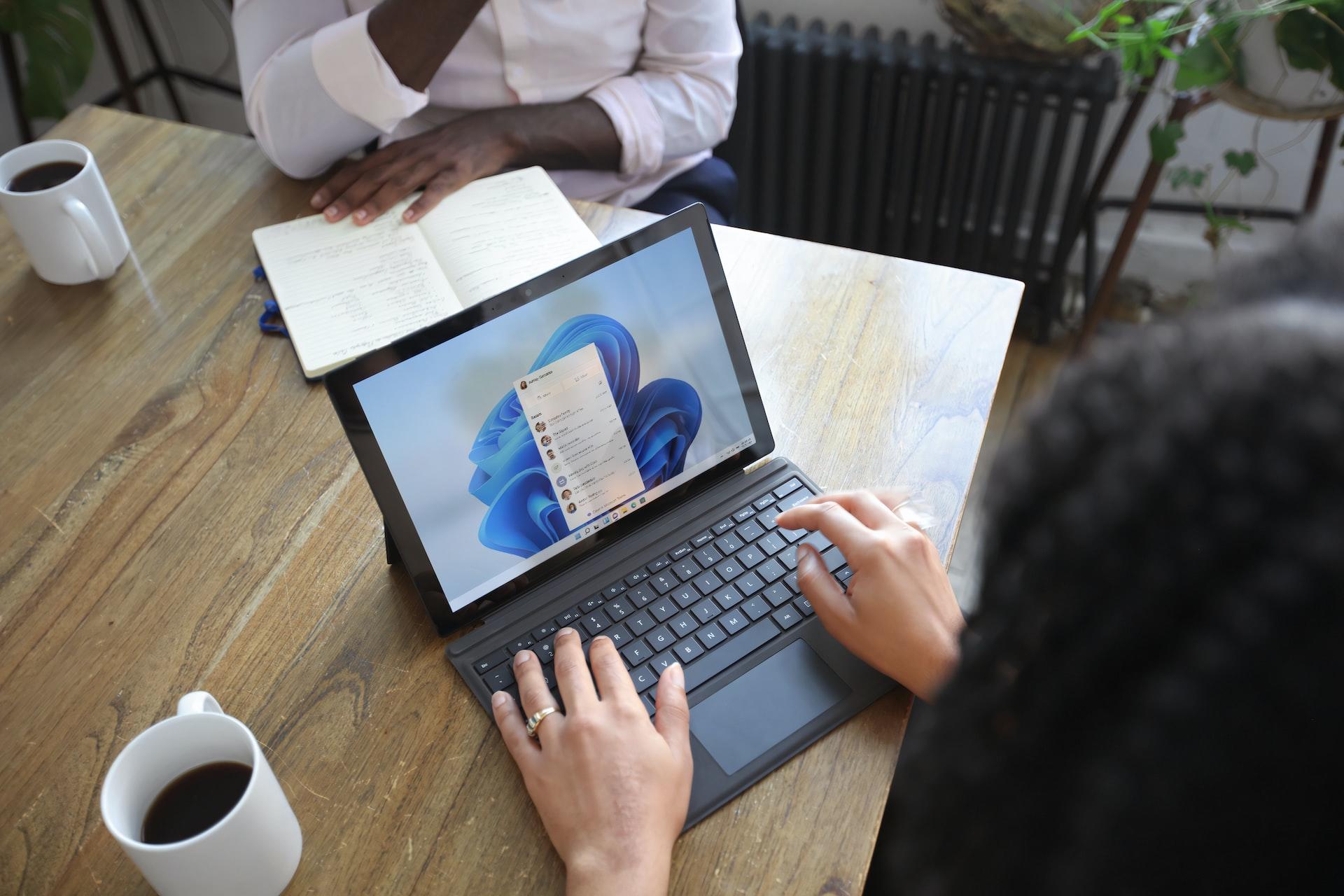 Eine Person sitzt an einem Tisch und hat auf einem Laptop das Startmenü geöffnet.