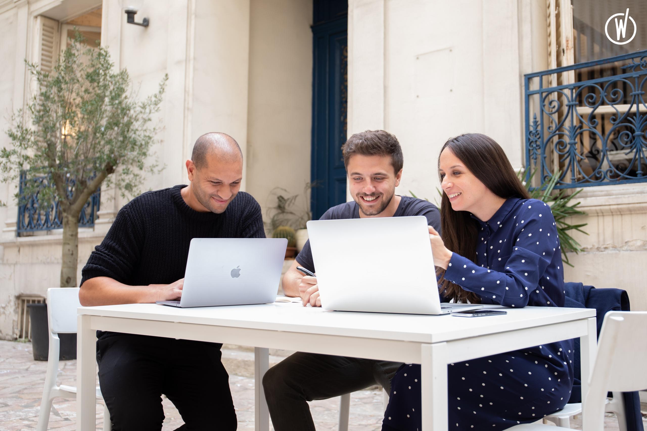 Drei Personen sitzen an einem Tisch und schauen lachend in zwei Laptops.