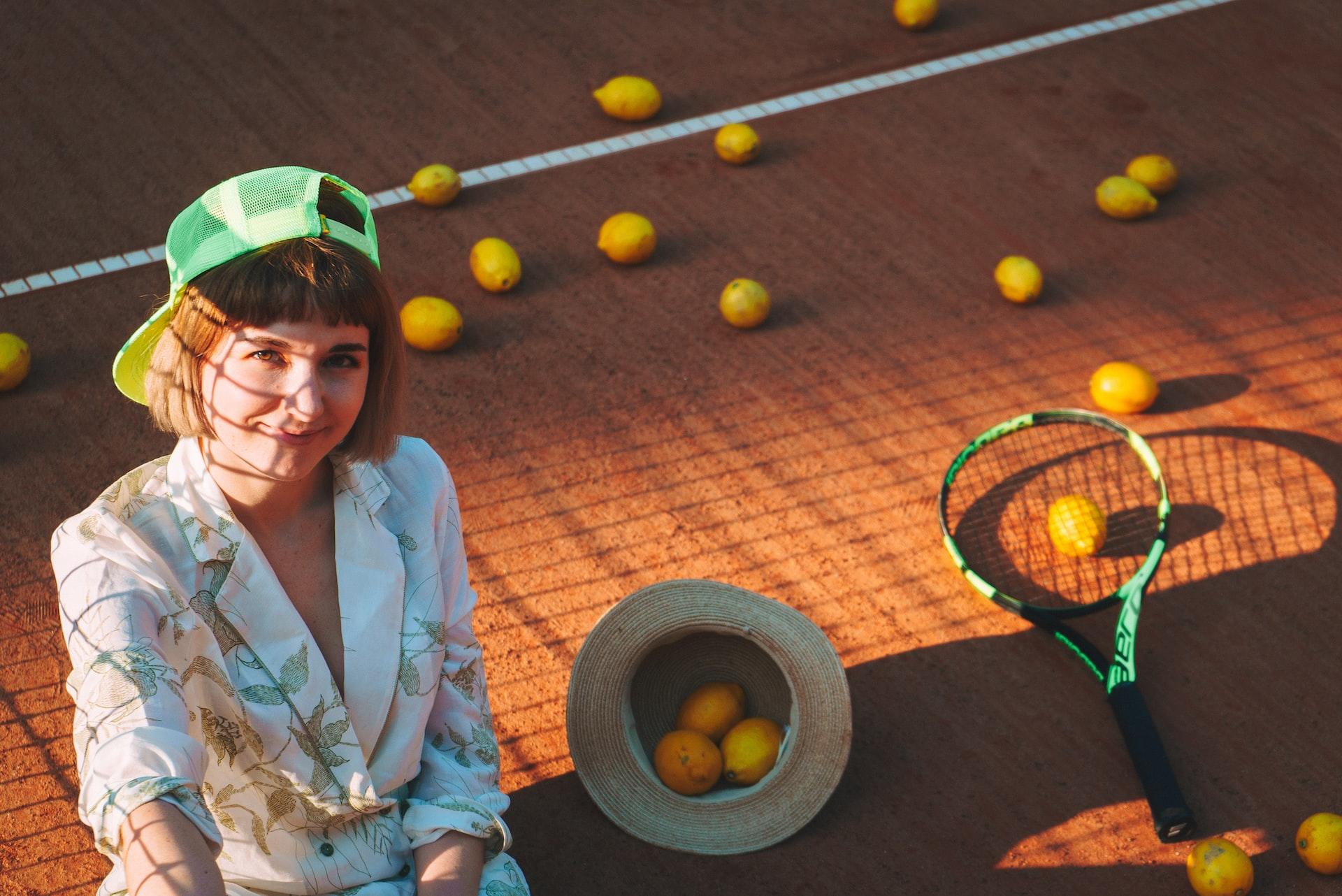 Eine Tennisspielerin mit Kappe steht auf dem Tennisplatz und neben dem Tennisschläger sowie in ihrem Hut liegen Zitronen anstatt Tennisbälle