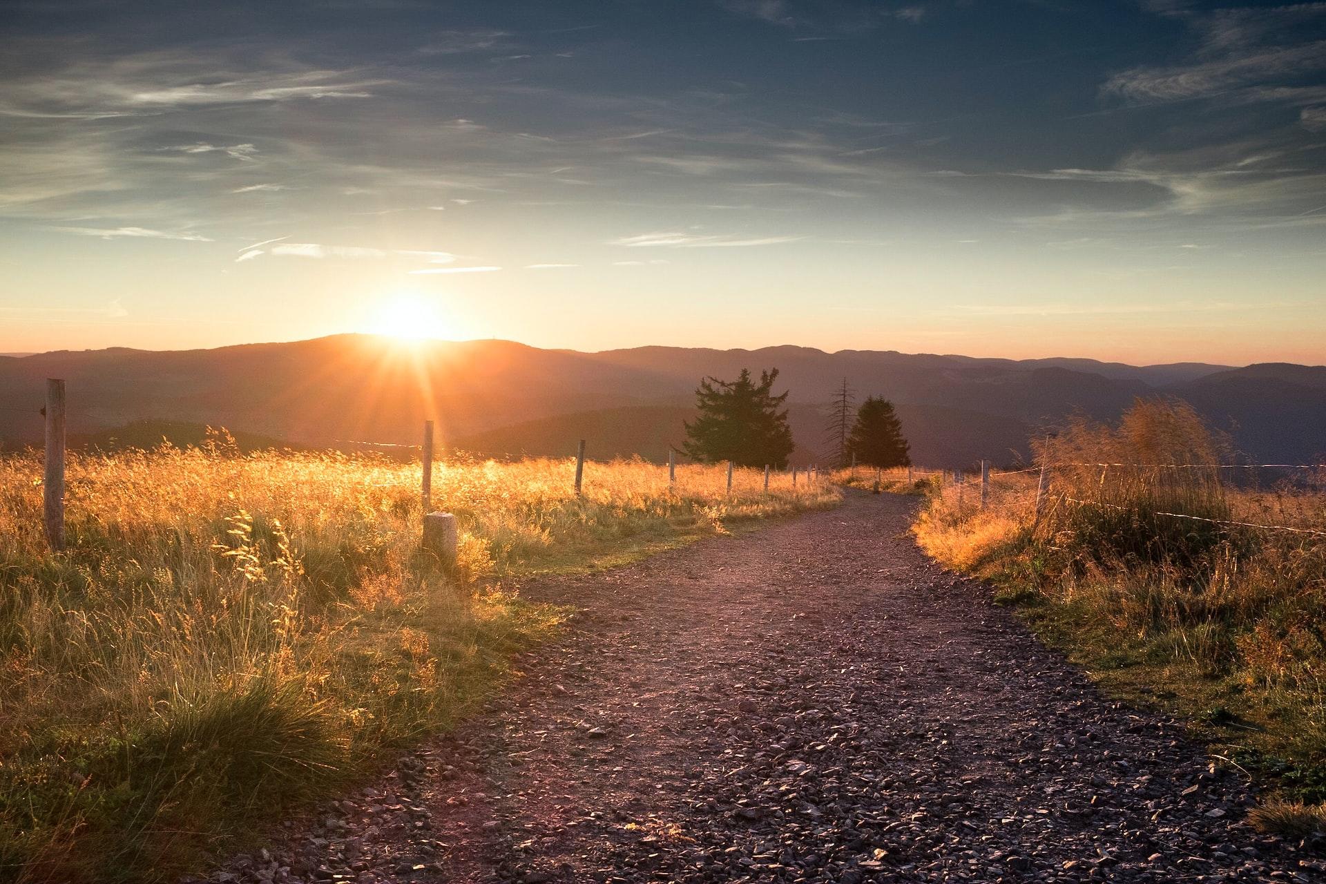Ein Weg durch ein Feld führt zu einem Tal über dessen Berge die Sonne untergeht.