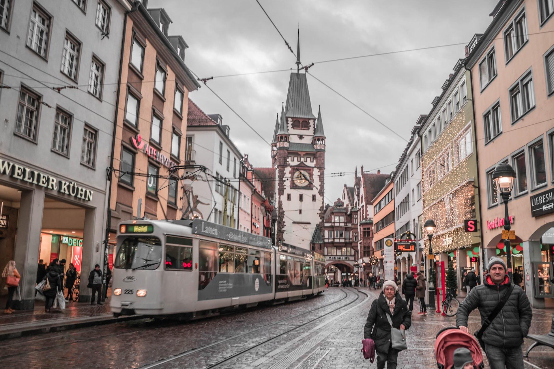 Menschen laufen durch die Fußgängerzone von Freiburg und eine Bahn fährt durch. 