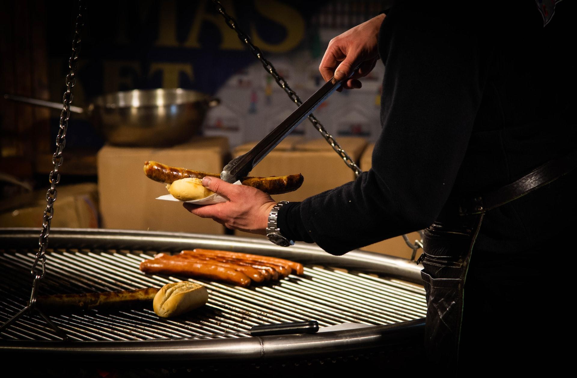 Ein Mann steht an einer Grill und legt eine Bratwurst in ein Brötchen.