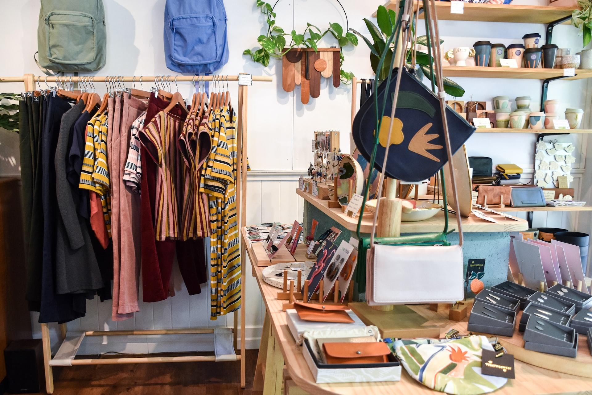 In einem kleinen Laden steht ein Kleiderständer und ein Tisch mit verschiedenen Taschen und Hüten.