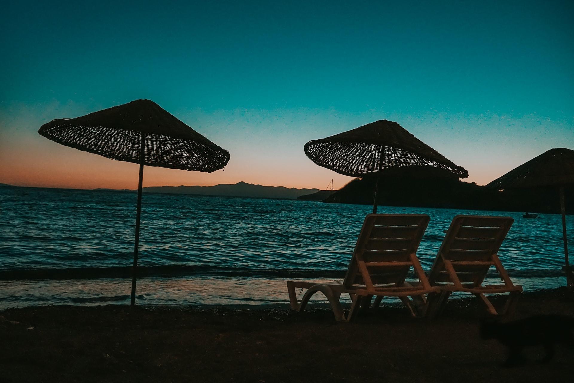 An einem Strand stehen zwei Liegestühle und zwei aufgespannte Sonnenschirme.