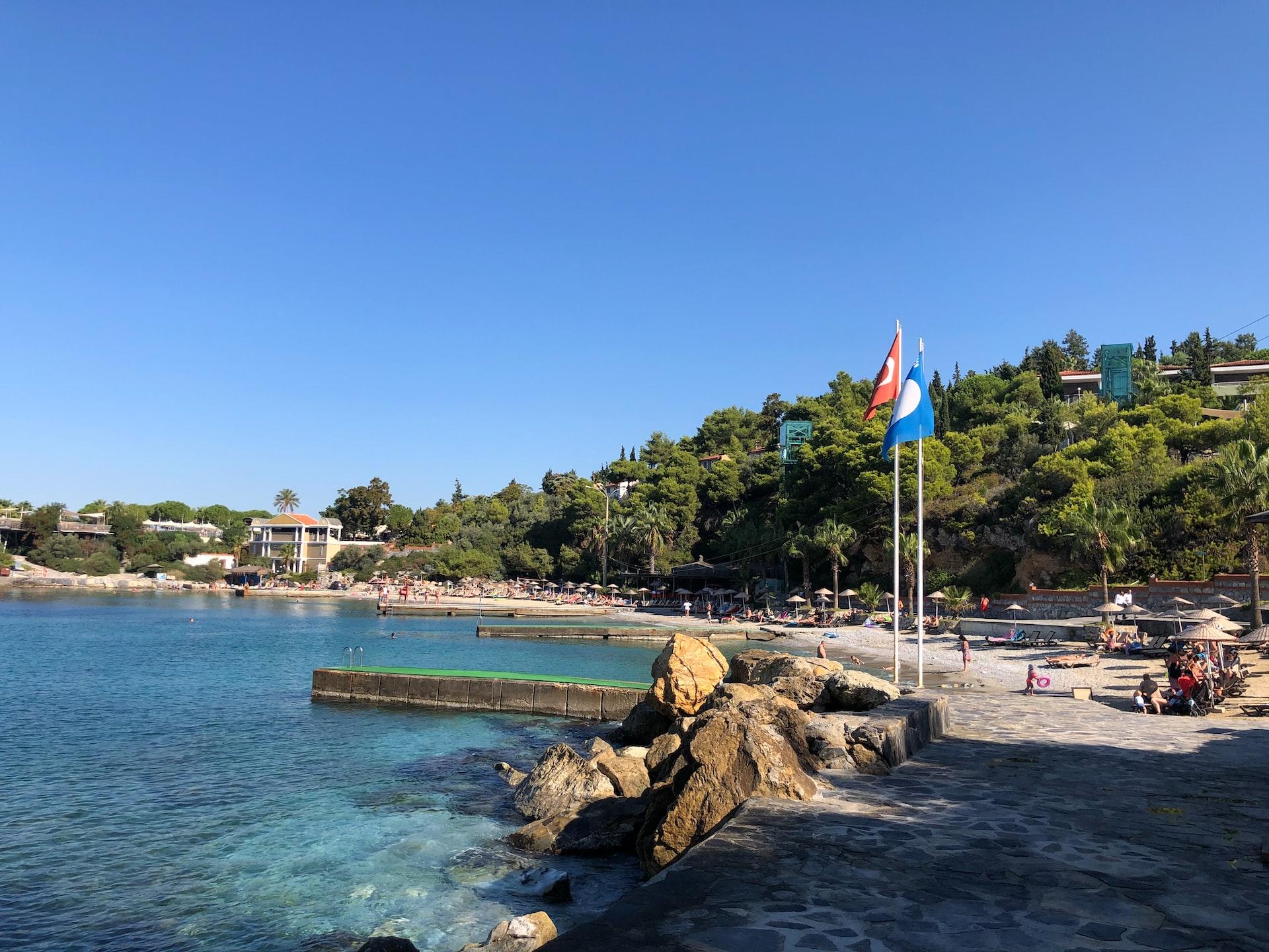 Vor einem Strand ragen Stege ins Wasser hinein und zwei Flaggen stehen auf einer Promenade.