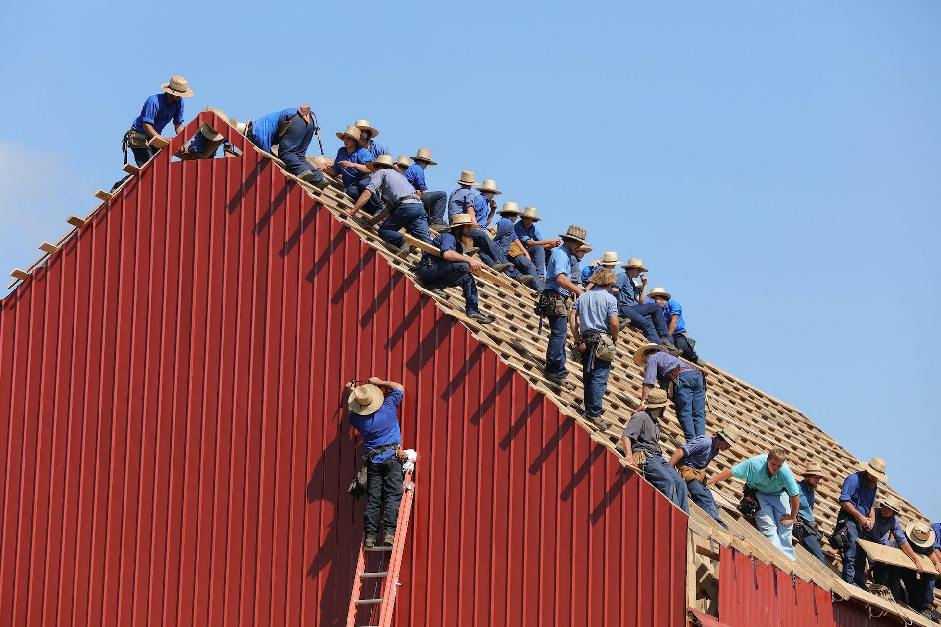 Männer in traditioneller Kleidung beim Dach decken eines rot gestrichenen Holzhauses.