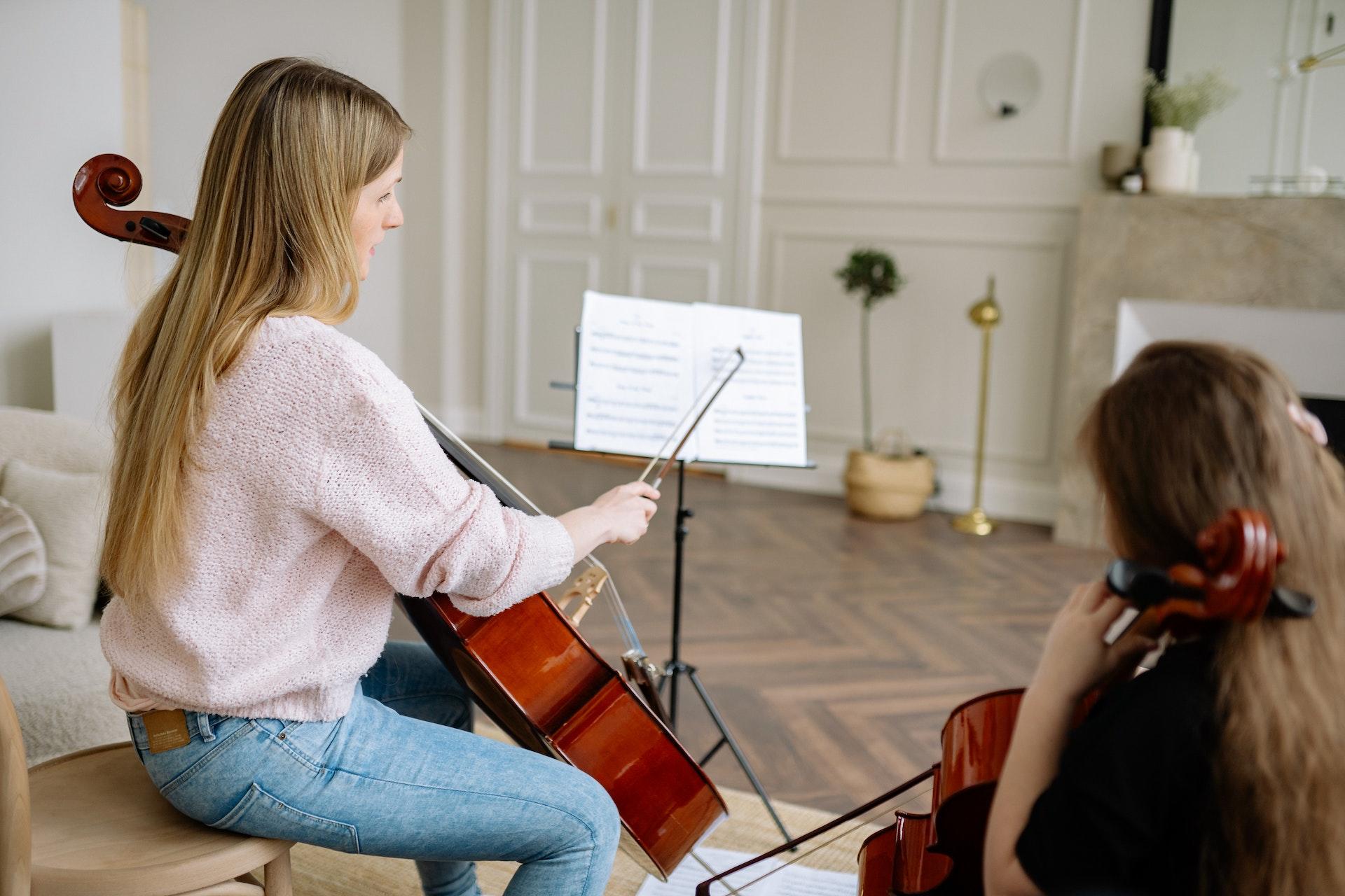 Zwei Personen halten je ein Cello und schauen auf ein Notenblatt auf einem Ständer.