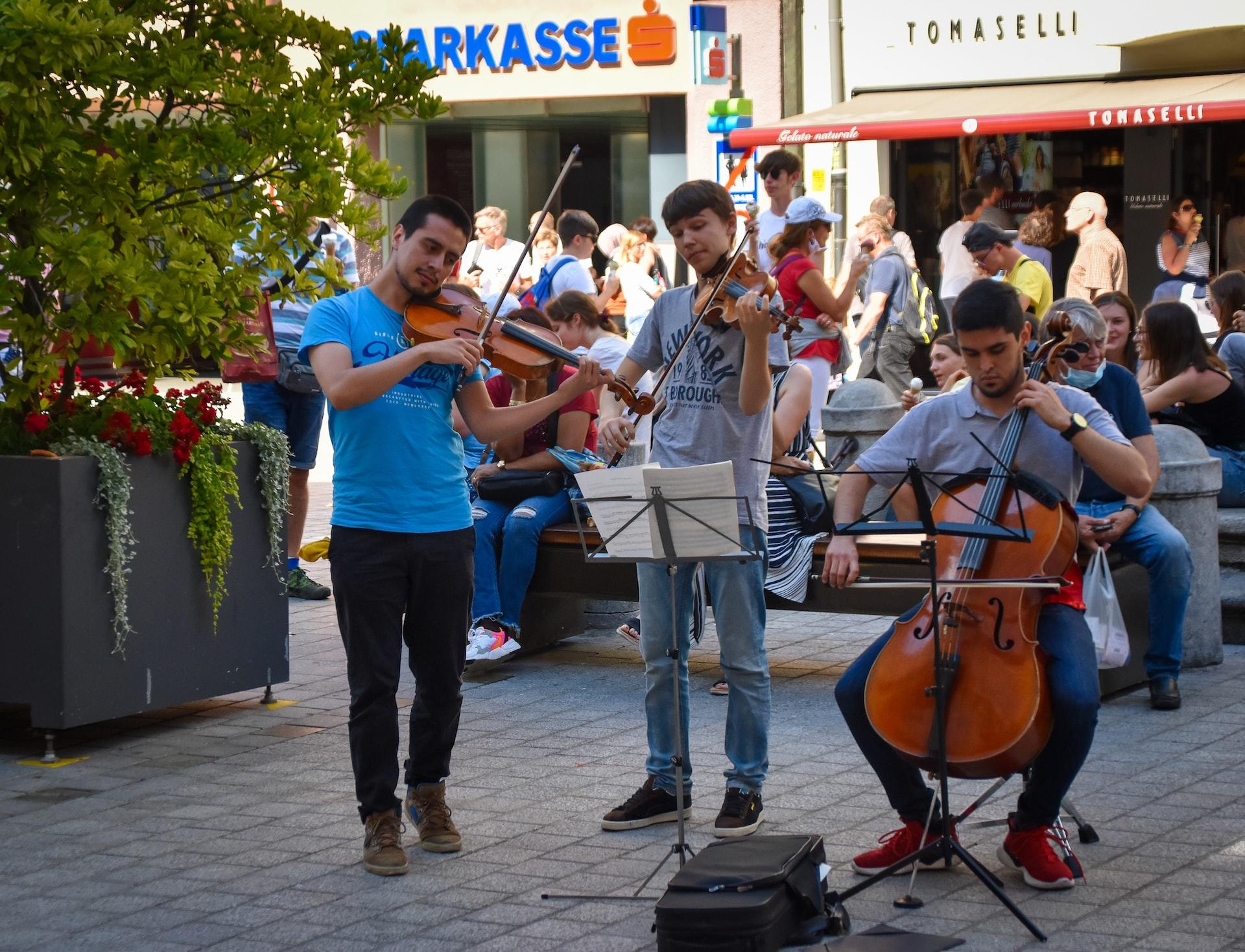 In einer städtischen Umgebung spielen zwei Personen stehend Geige und eine Person sitzt am Cello.