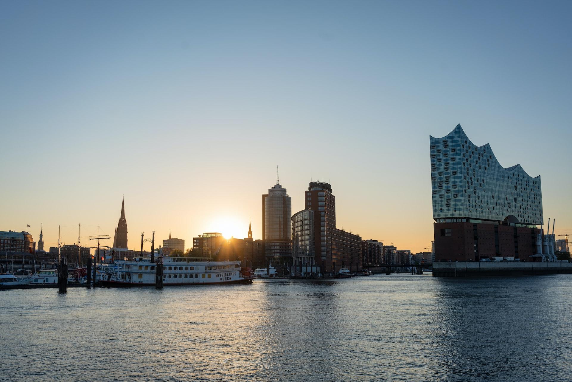 Hinter modernen Gebäuden am Wasser leuchtet die untergehende Sonne hell.