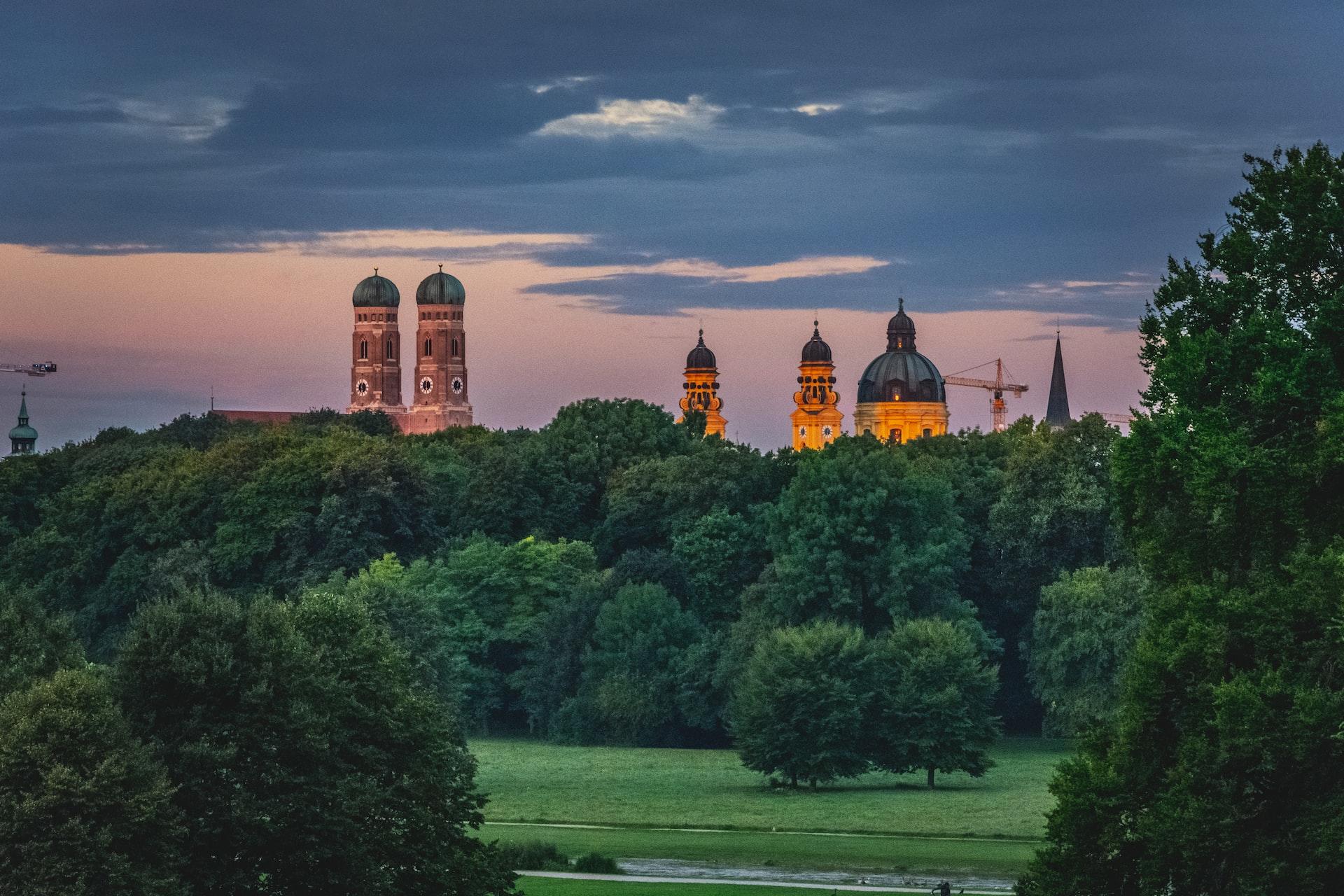 Hinter hohen Bäumen und Büschen ragen die Türme und Kuppeln von alten Gebäuden in den Himmel.