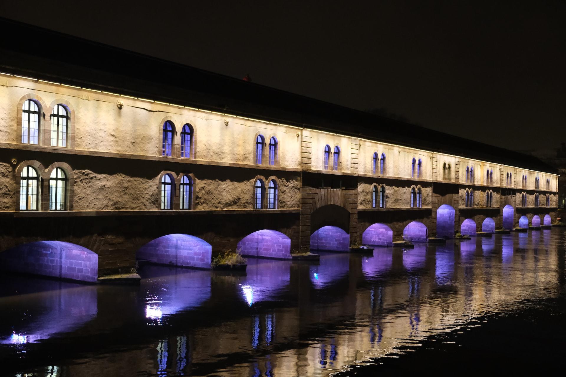Eine alte Brücke in einer elsässischen Stadt bei Nacht mit einer Wasserspiegelung der lila beleuchteten Brückenbögen.