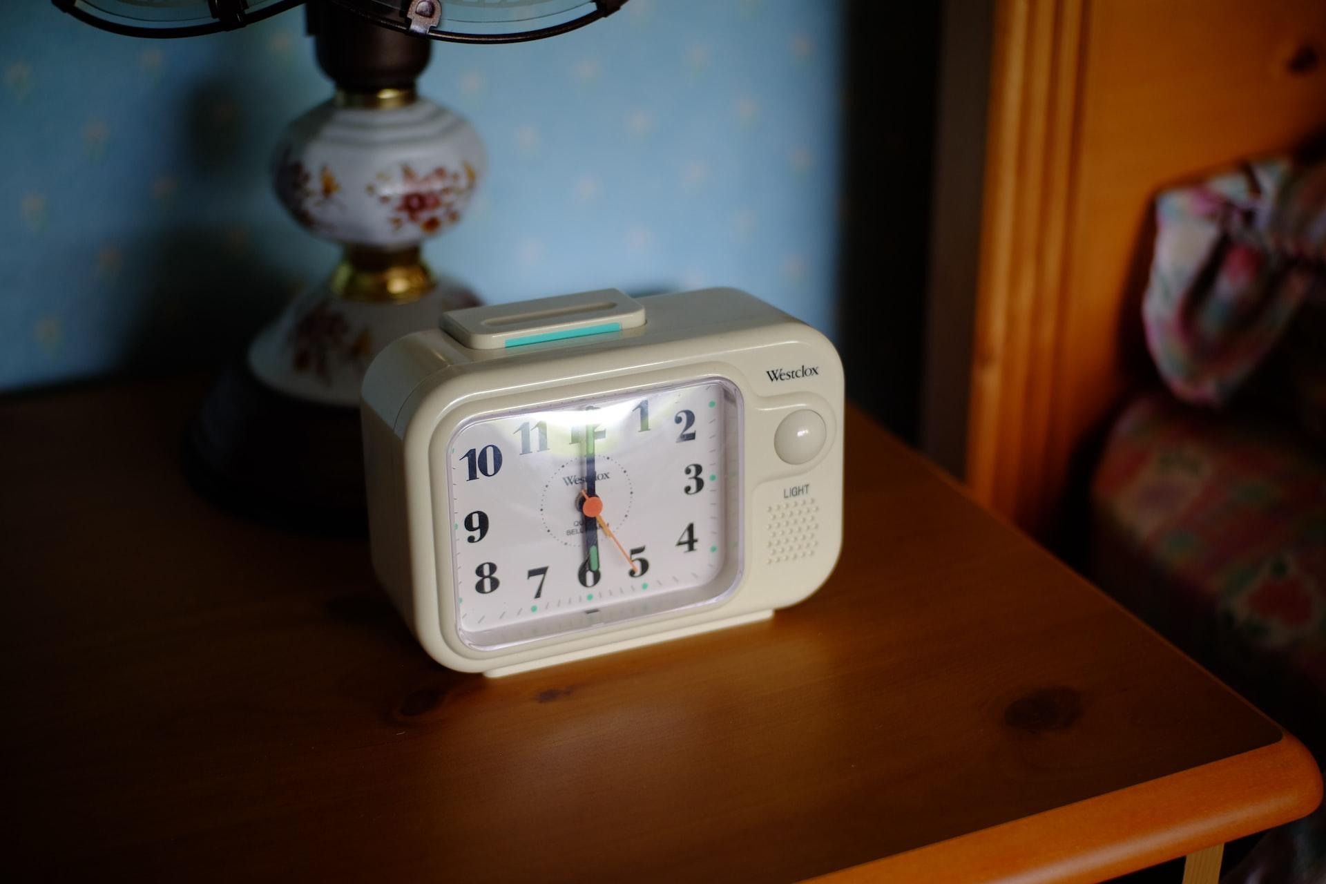 Auf einem Nachttisch steht ein analoger Wecker und die Zeiger stehen auf sechs Uhr.