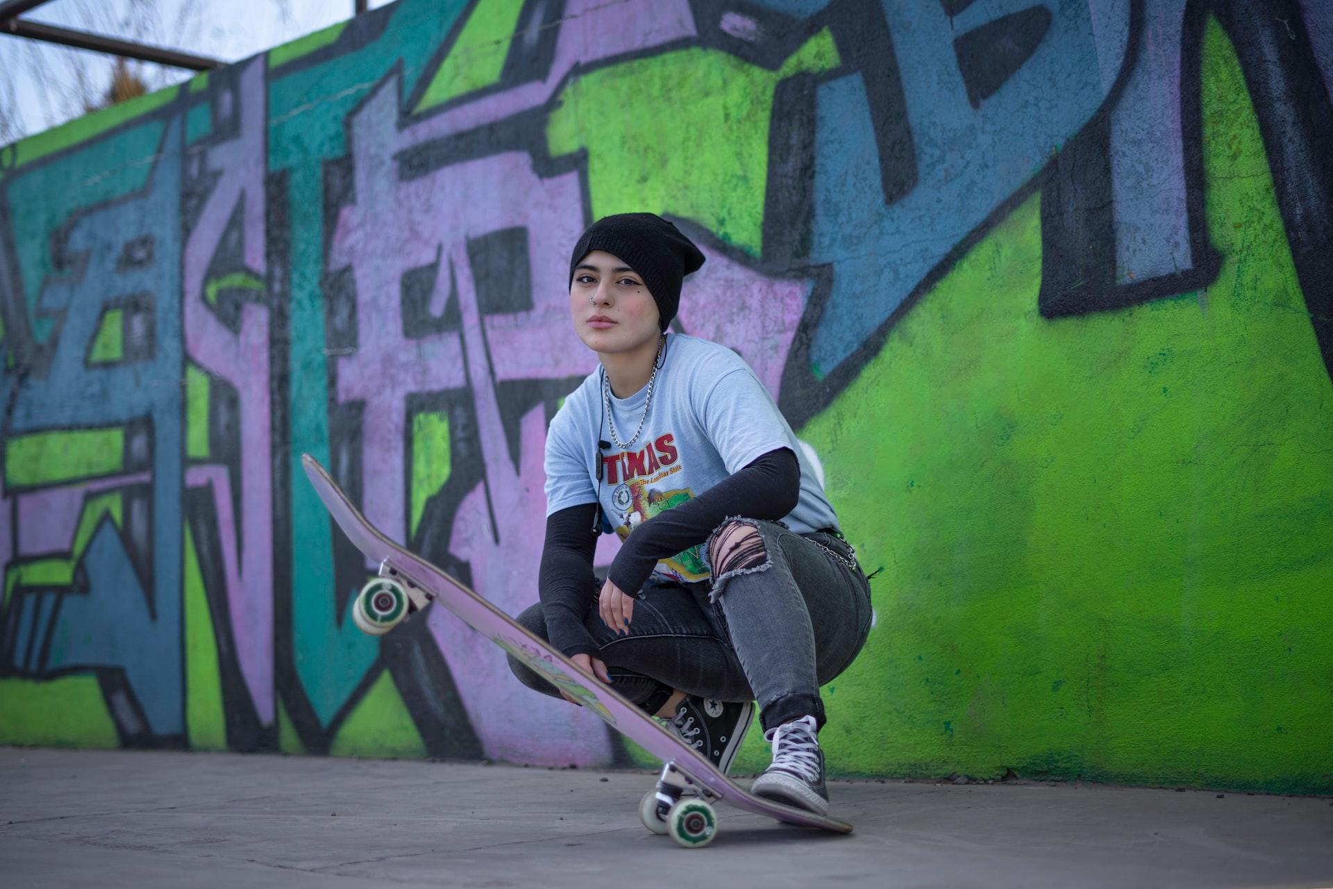 Eine Person mit Mütze und Skateboard hockt vor einer mit Graffiti besprühten Wand.